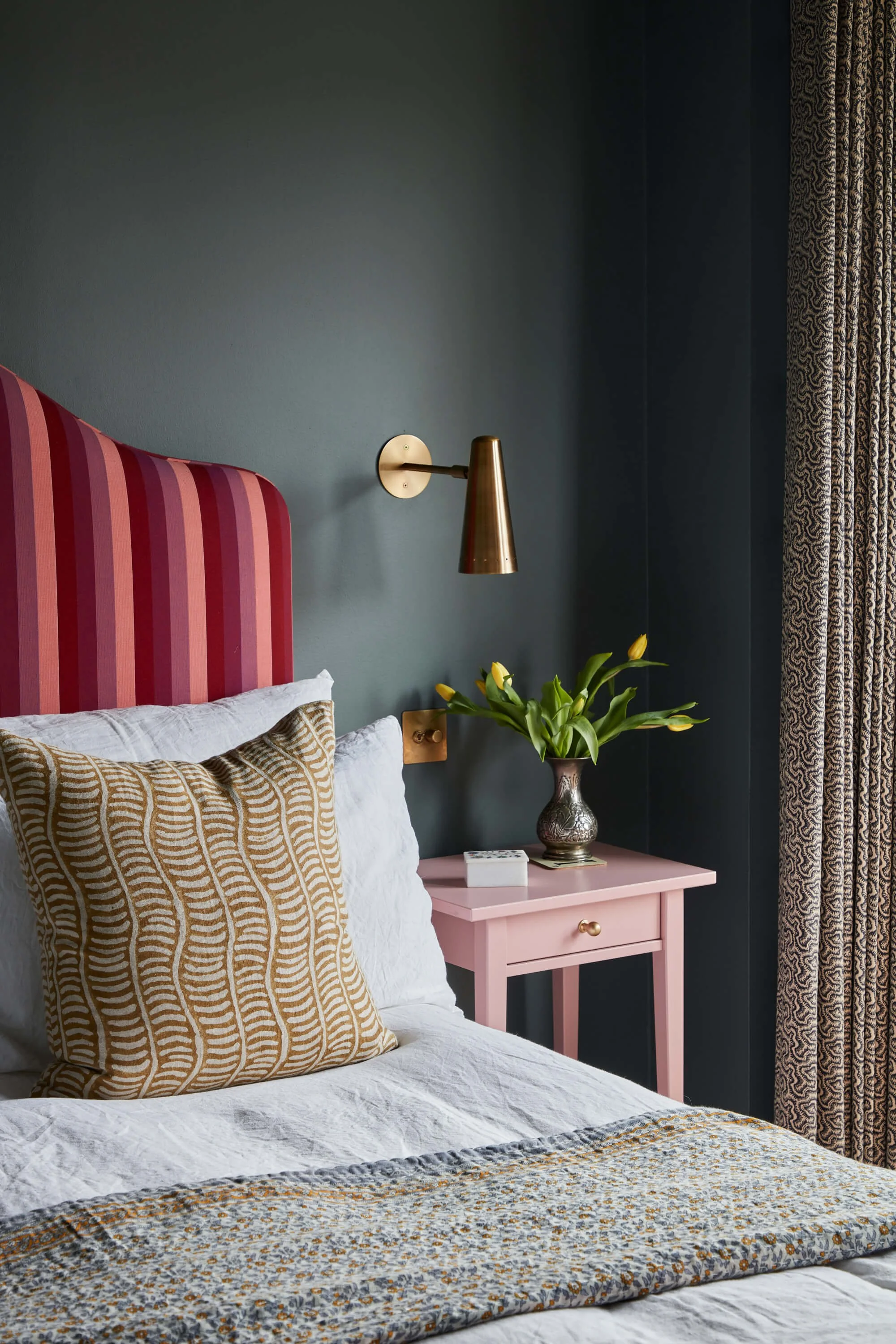 A cozy bedroom with a bed that has a tall pink and red striped headboard, white bedding, and decorative pillows with curved line patterns. Next to the bed is a pink nightstand with a brass lamp, a vase filled with yellow tulips, and a small white box. The wall behind the bed is painted dark blue, with patterned curtains drawn to the side.