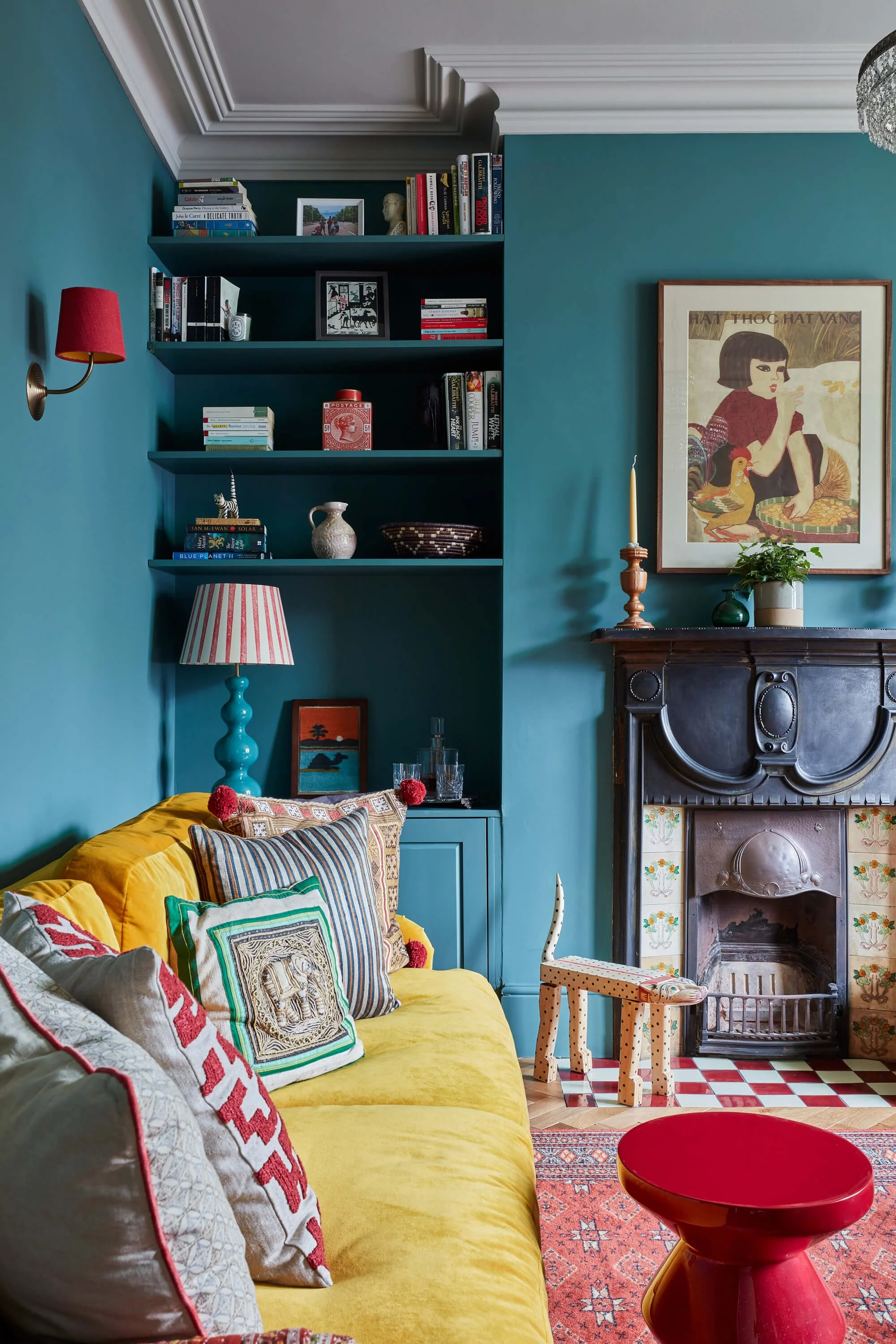 A colorful living room with a yellow sofa decorated with pillows, a blue lamp, a fireplace with ornate tiles, and a bookshelf against teal walls. There is a framed artwork, a potted plant, a candlestick, and a small polka-dotted stool.