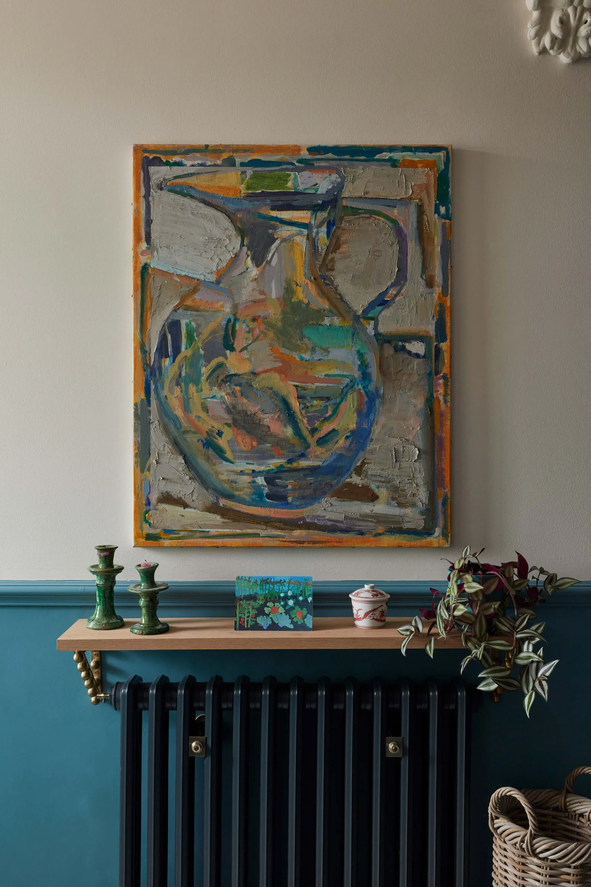 Decorative display with green candle holders on a wooden table, a small colorful painting, a white and red ceramic jar, and a potted plant with dark purple and green leaves, against a two-toned wall with blue at the bottom and beige above, topped with a large abstract painting.