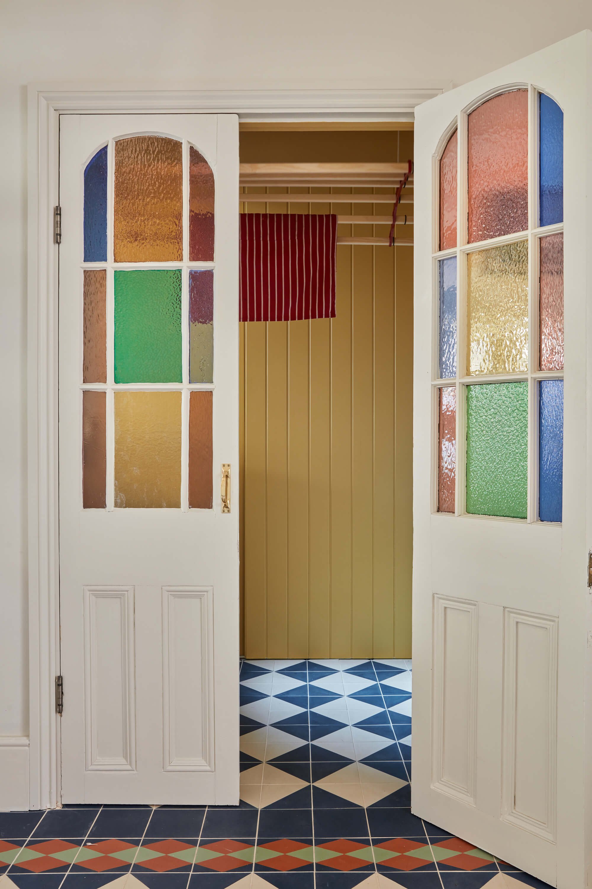 Open white double doors with textured, multi-colored glass panels, revealing a yellow-walled room with a wooden shelf and a red and white striped towel hanging.