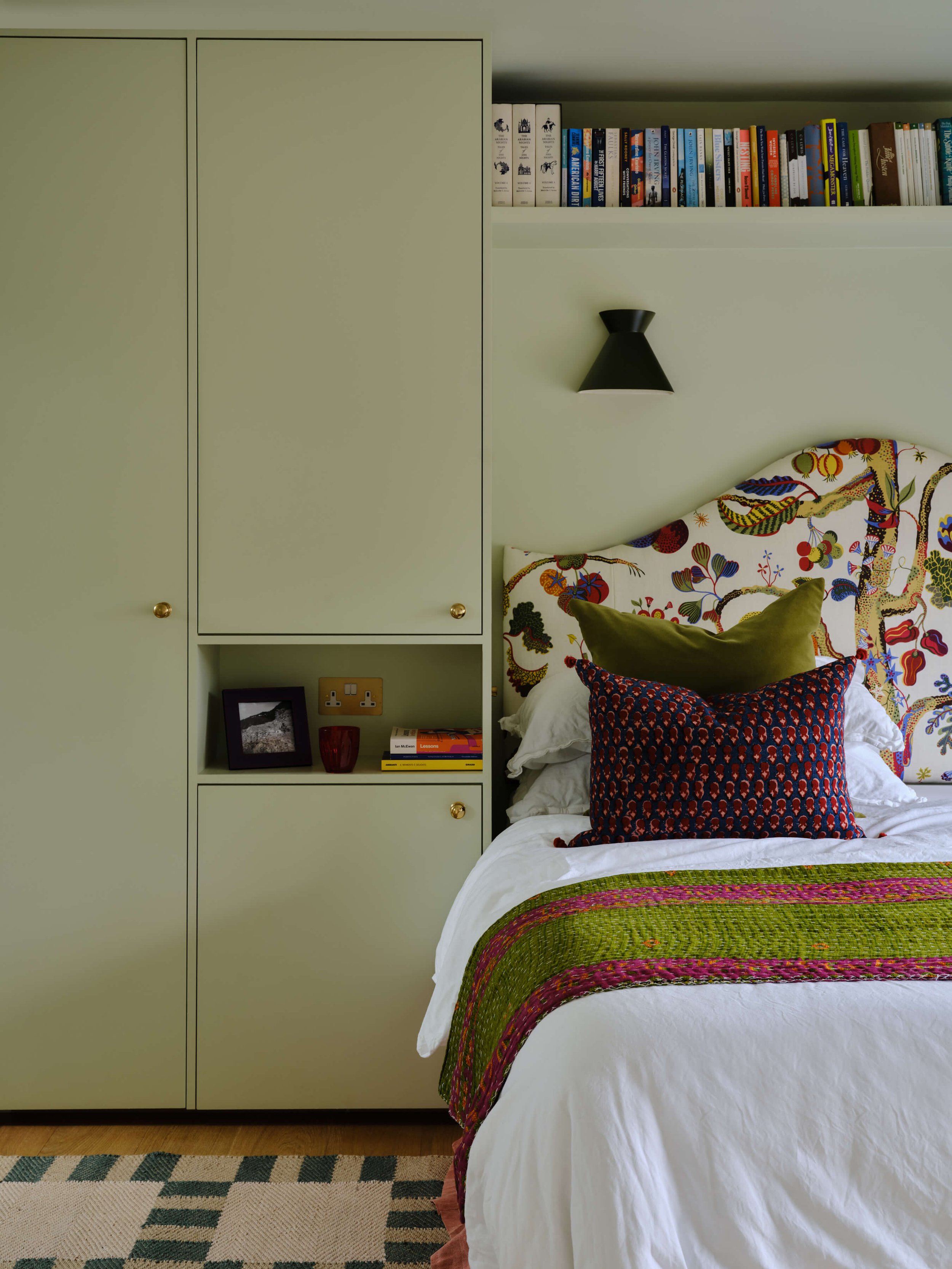 Bedroom with a colourful floral upholstered headboard, built-in sage green wardrobes, a black wall light and a geometric patterned rug.