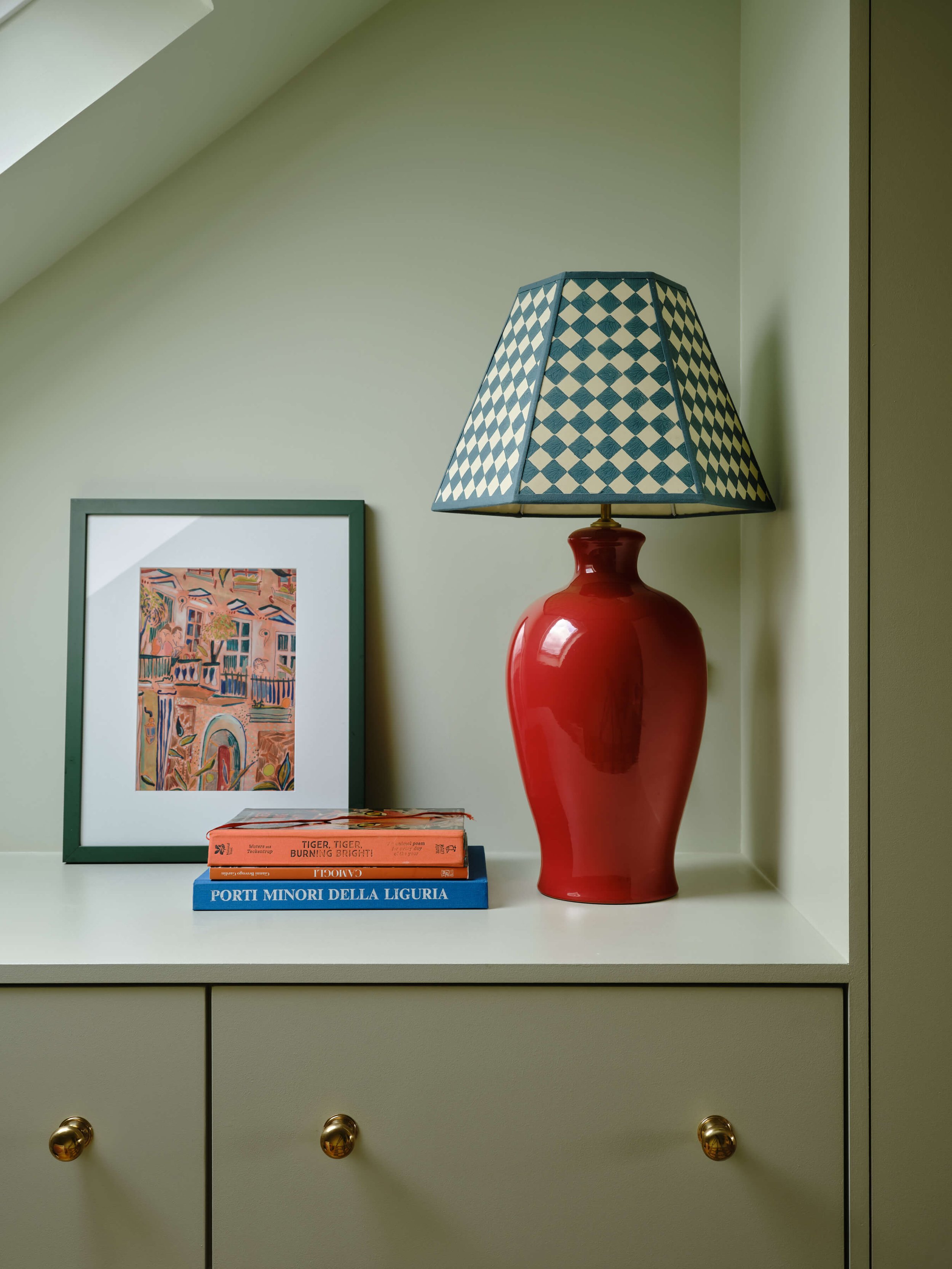 Green chest of drawers with brass knobs, styled with a red ceramic lamp, gingham lampshade, framed abstract print and stacked books.