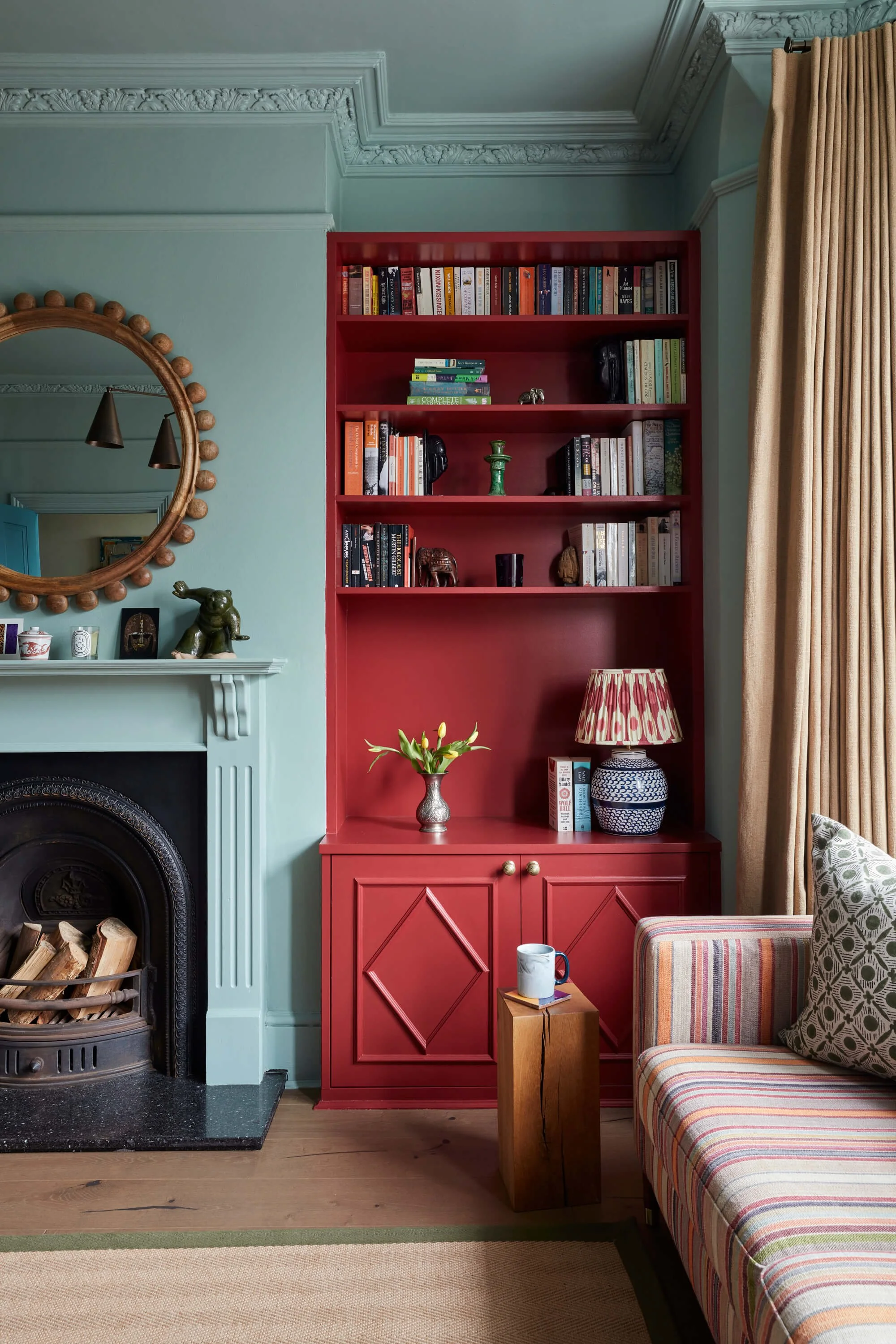 A living room with a green fireplace, a round mirror with wooden beads on the frame above it, a white mantel with small decorative items, a red bookshelf filled with books and small decor, a beige curtain on the right, a table lamp with a patterned shade and a large striped armchair with a patterned cushion, a small wooden side table with a coffee mug, and a beige rug with a green stripe at the bottom.
