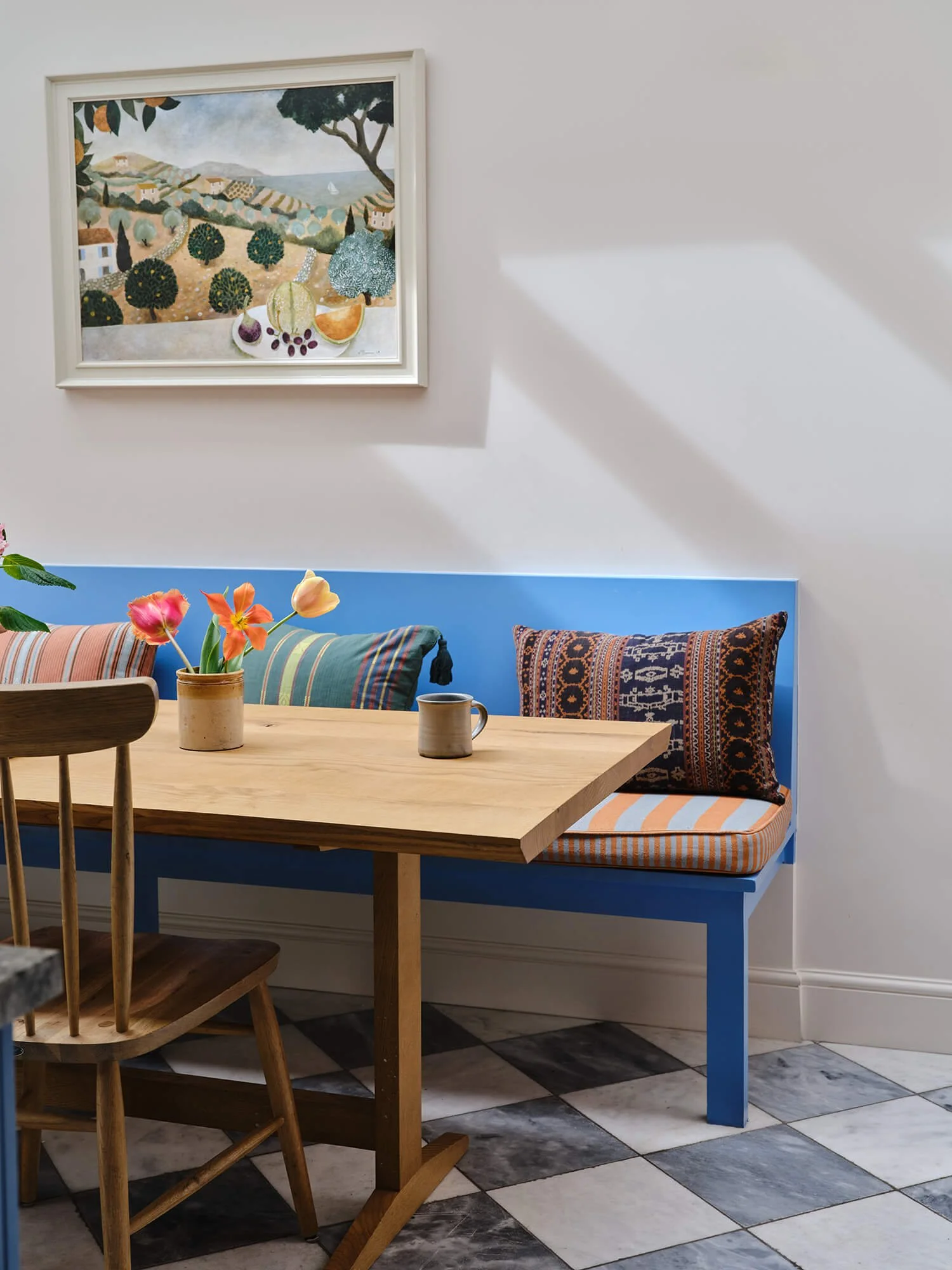 Interior of a dining area with a wooden table, two chairs, and a built-in seating nook with blue and patterned cushions. A small potted plant with pink and orange tulips is on the table. A framed painting of a countryside landscape hangs on the white wall, with sunlight casting a shadow on the wall.