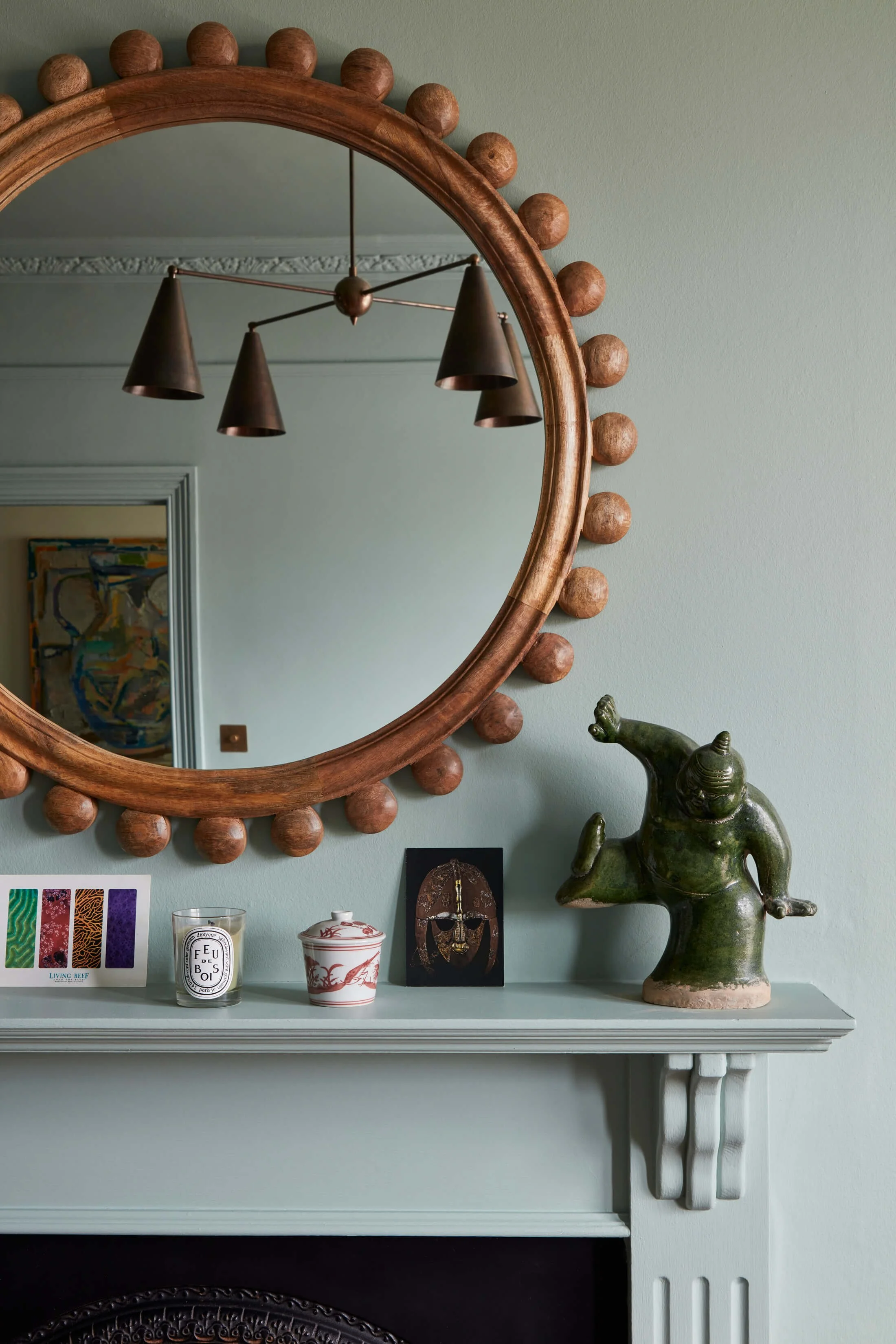A wall mirror with a wooden frame decorated with round wooden bumps surrounds a room with art and decorative objects. Below the mirror, a mantel displays a colorful card, a candle, a porcelain cup and saucer, and a black artwork of a face. To the right, a green ceramic sculpture of an elephant stands on the mantel.