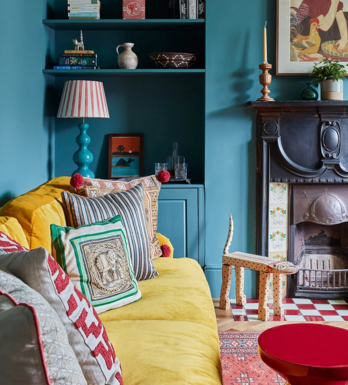A colorful living room with a bright yellow couch adorned with decorative pillows, a tall turquoise lamp with a pink and white striped lampshade, and a wooden fireplace mantel decorated with a candle, potted plant, and framed artwork. There are built-in shelves with books, vases, and decorative items. A small wooden animal sculpture with a long neck stands near the fireplace, and the room has a mix of patterns and vibrant colors.