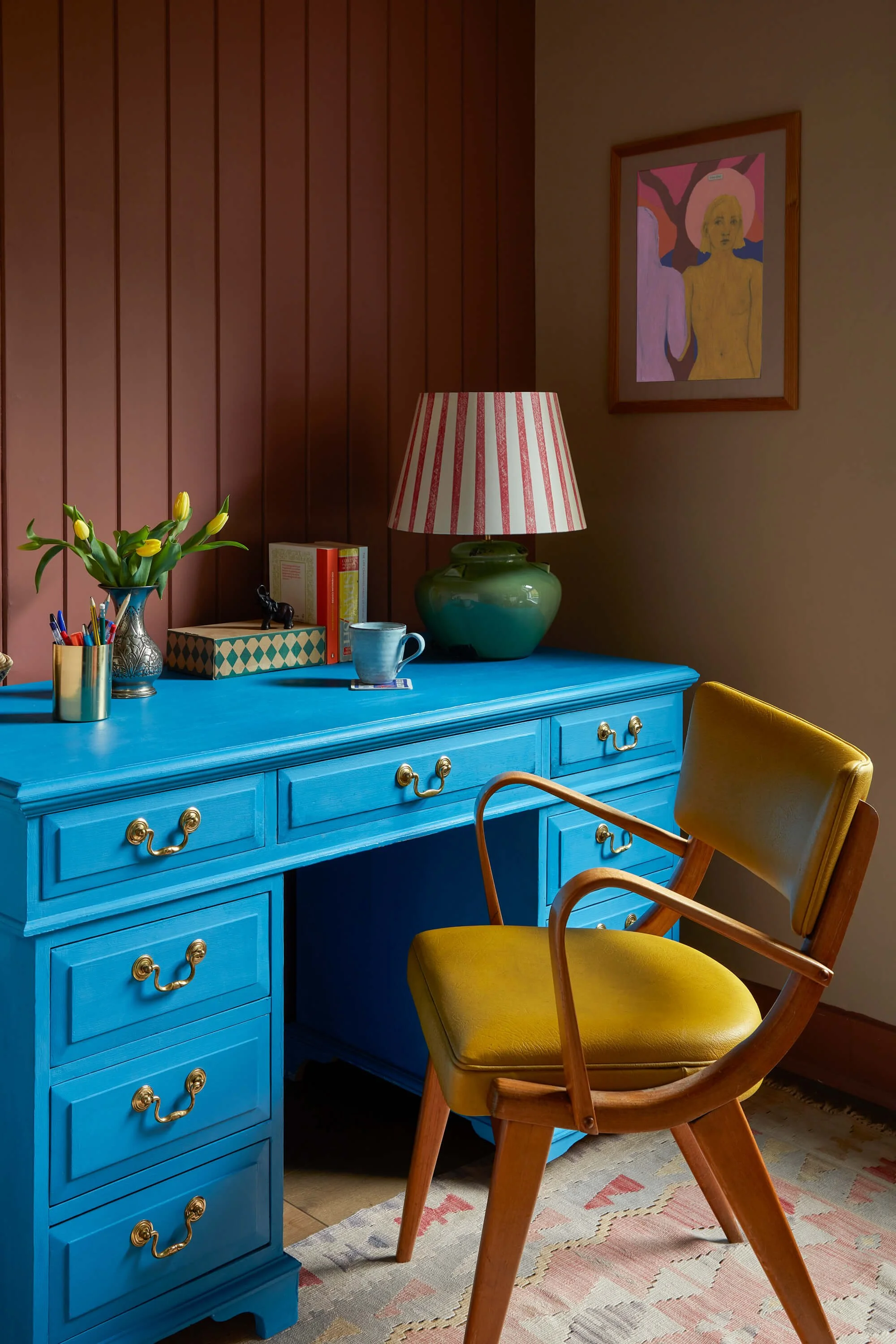 A blue dresser with gold drawer pulls on a wooden floor alongside a yellow chair with a wooden frame. The dresser holds a ceramic vase with yellow tulips, a box, a small black dog figurine, books, a coffee mug, and a lamp with a red and white striped shade. Behind the dresser, a wall features a framed artwork of two abstract women with a pink background. A beige and pink patterned rug covers part of the floor.