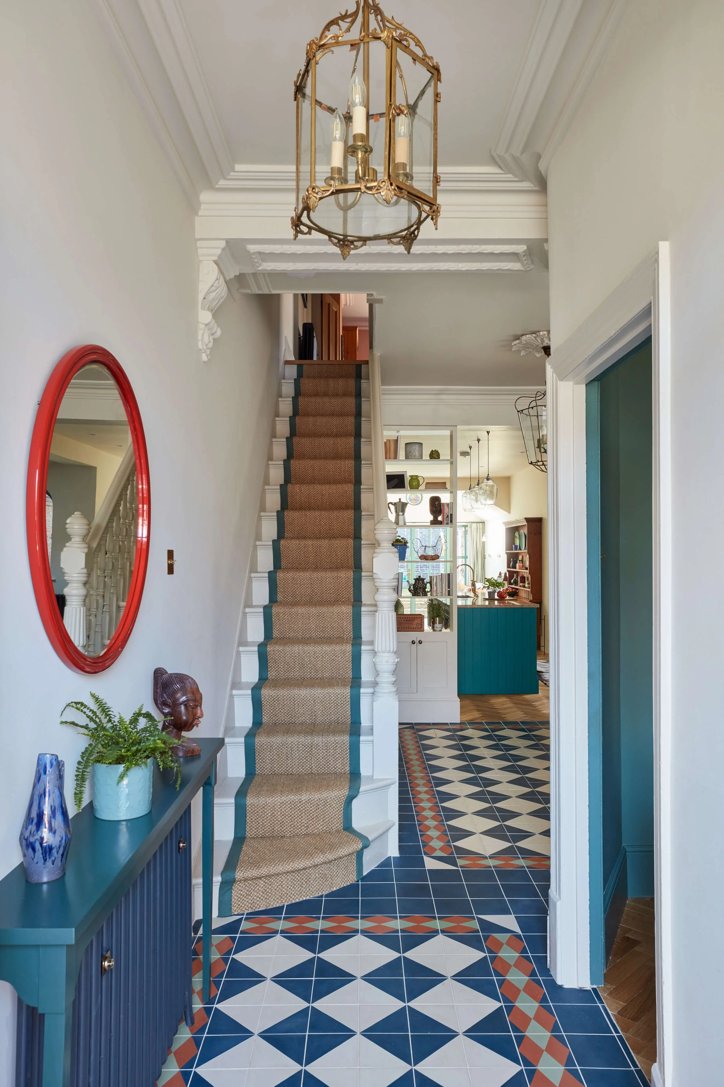 Entryway with a staircase and an ornate brass and glass chandelier hanging from the ceiling. To the left, a blue console table with decorative vases and a plant. The staircase has a beige runner with green borders. The checkered tile floor features blue, white, and red geometric patterns leading into a dining area with open shelving and a kitchen in the background.