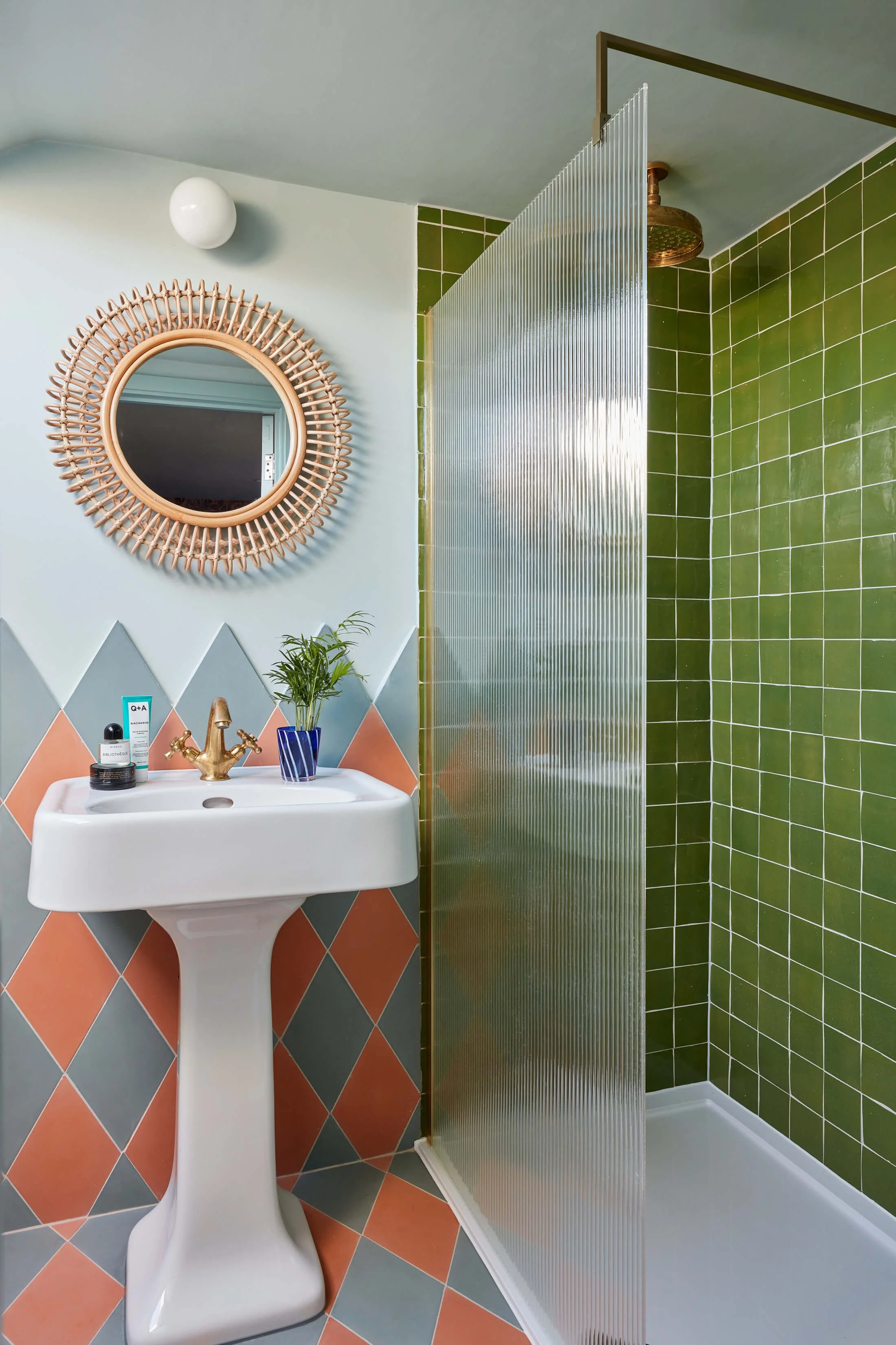 A bathroom with a white sink, a gold faucet, a round mirror with a woven rattan frame, green tiled shower area with a metallic divider, and pink and gray diamond-patterned floor tiles.