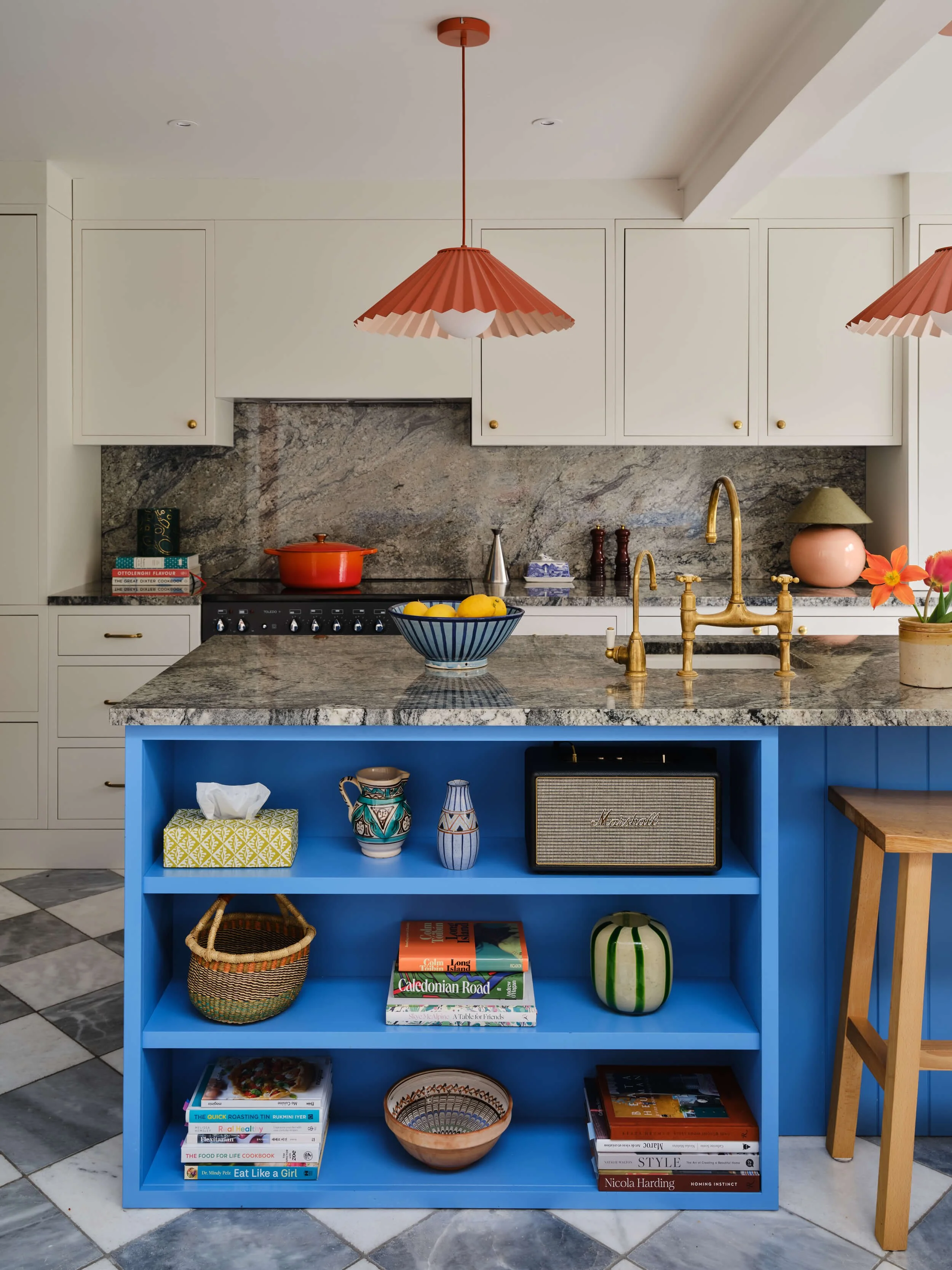 A kitchen island painted bright blue with shelves containing books, decorative vases, a basket, and a bowl. A woven basket is on the floor in front. The kitchen has white cabinets, a granite countertop, and a gold faucet. There are two coral-colored pendant lights and a small wooden stool.