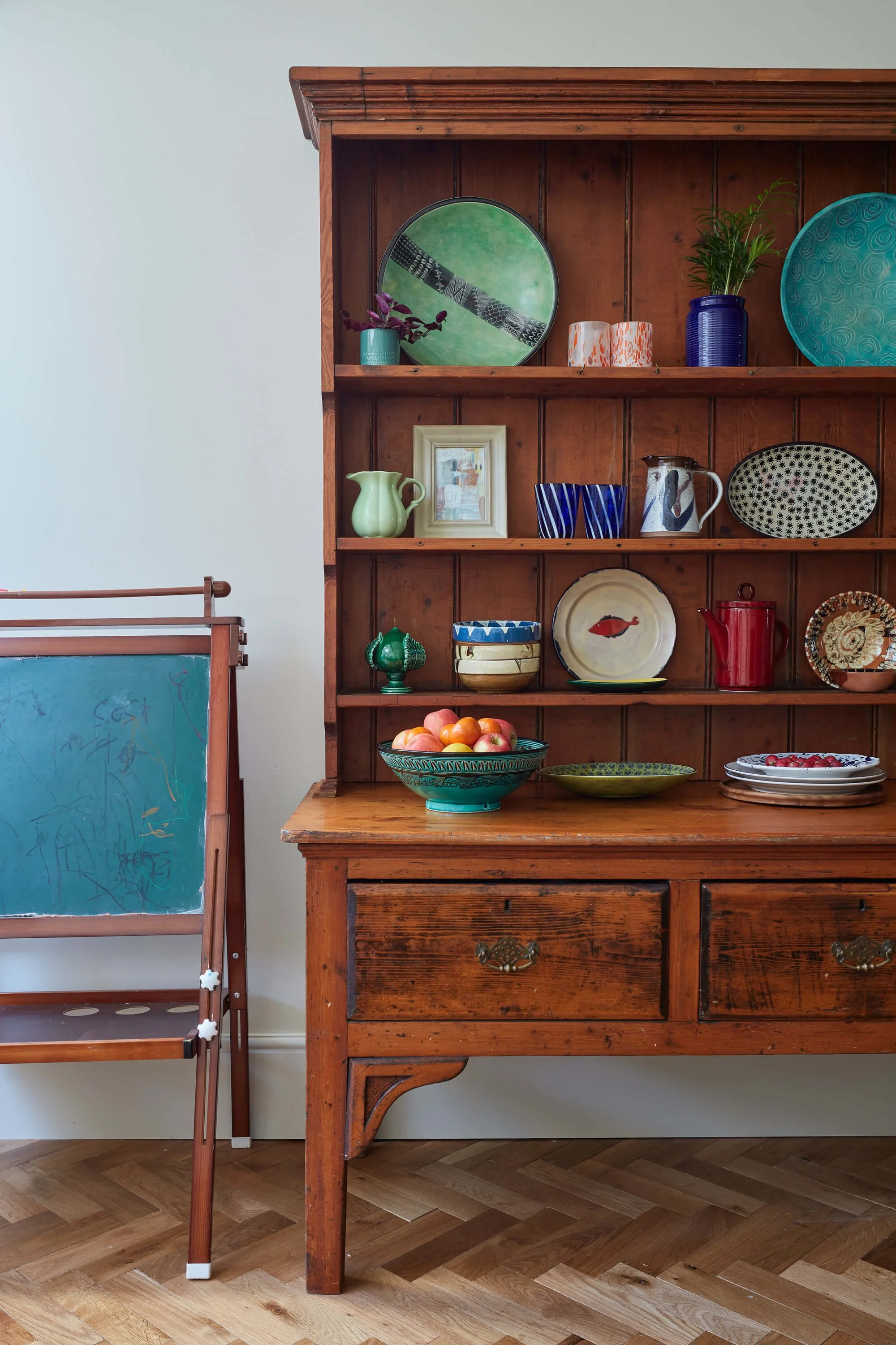 A wooden display shelf with various decorative items, including colorful vases, a small framed picture, and ceramic plates, with a green bowl of apples on the desk below. To the left, a chalkboard easel with a blue surface is visible.