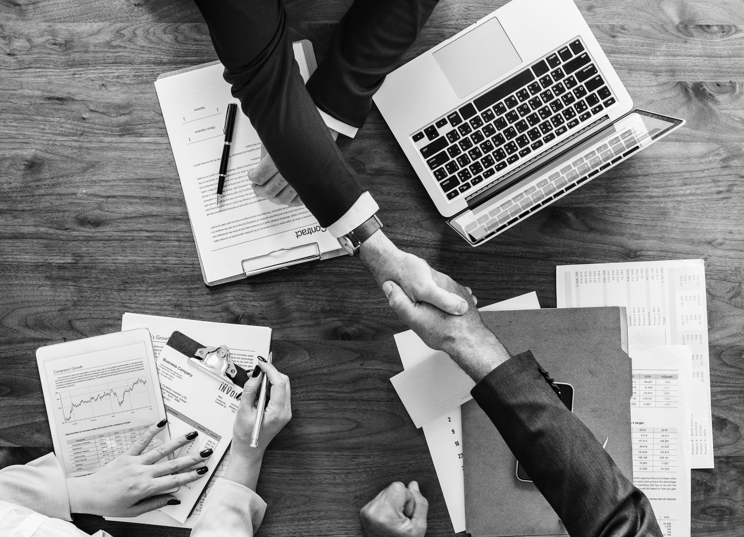 Reunião de negócios com pessoas trocando um aperto de mãos ao redor de uma mesa com laptops, documentos, gráficos e planilhas.
