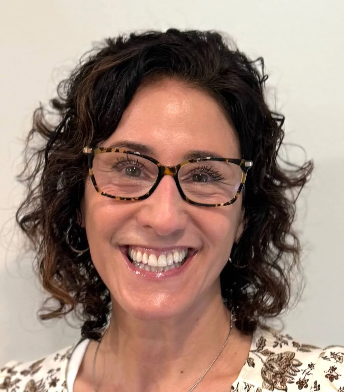 Karen Green, founder of Conscious Care Cards, smiling close-up with curly dark hair and tortoiseshell glasses, wearing a white floral shirt, representing wellness and mindfulness.