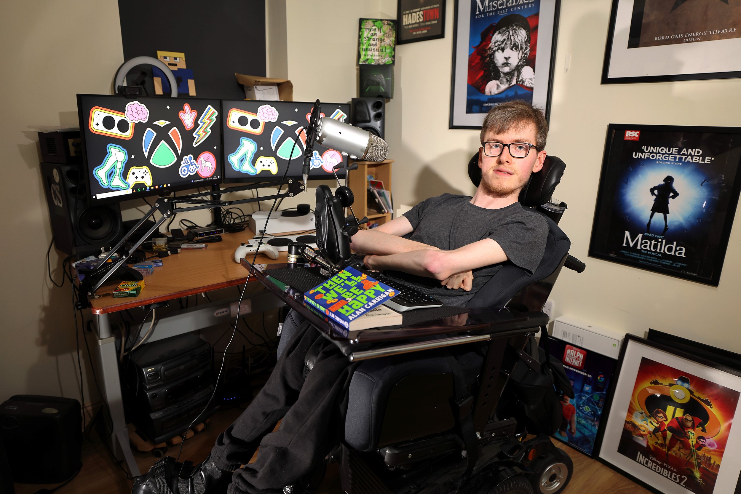 A man (Alan Carrigan) sitting in a motorised wheelchair with his arms crossed and his book in front of him.