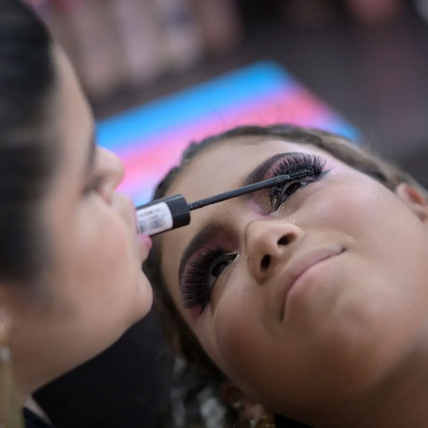 A women (Clélia Rodriguesff) holding a mascara wand in her mouth, applying the product on a woman's lashes