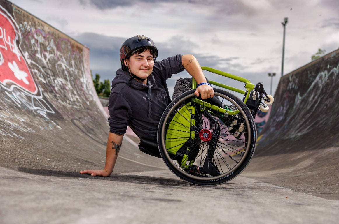 A man (Milo Watson) doing a wheelchair trick in a skate park.