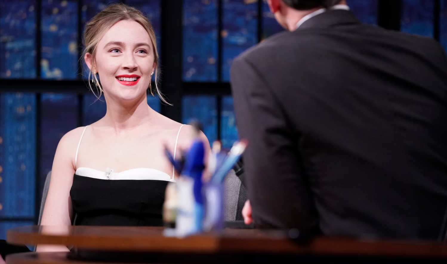 A woman (Saoirse Ronan) wears a black dress and red lipstick, and is smiling as she speaks.