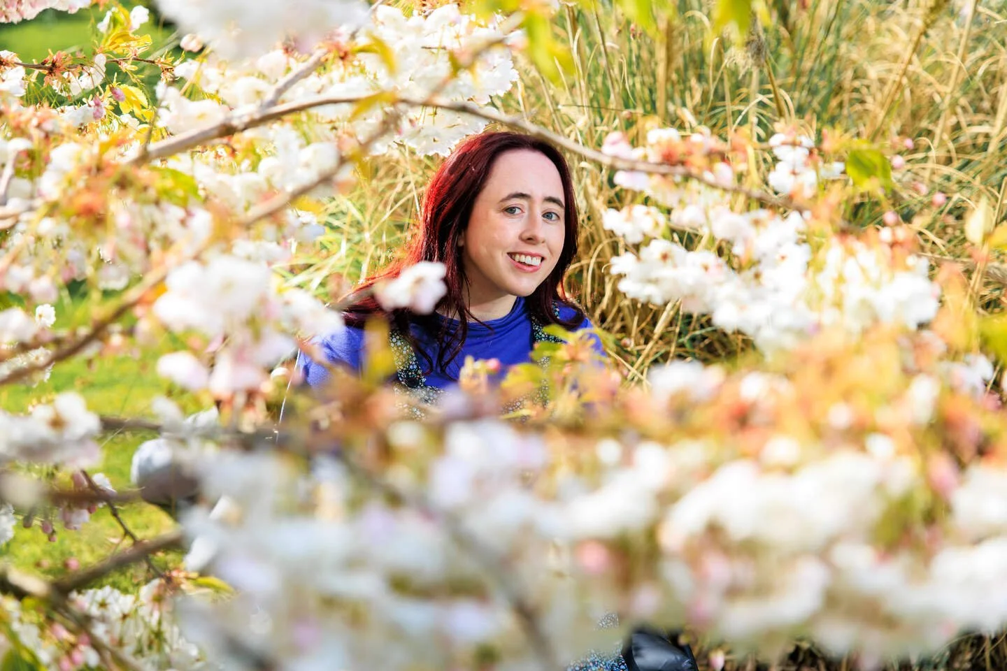 A woman (Niamh Ní Hoireabhaird) smiling at the camera, surrounded by flowers.