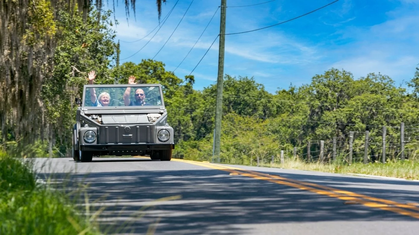 Vintage open-top Volkswagen driving along a sunny, tree-lined road with two passengers inside.