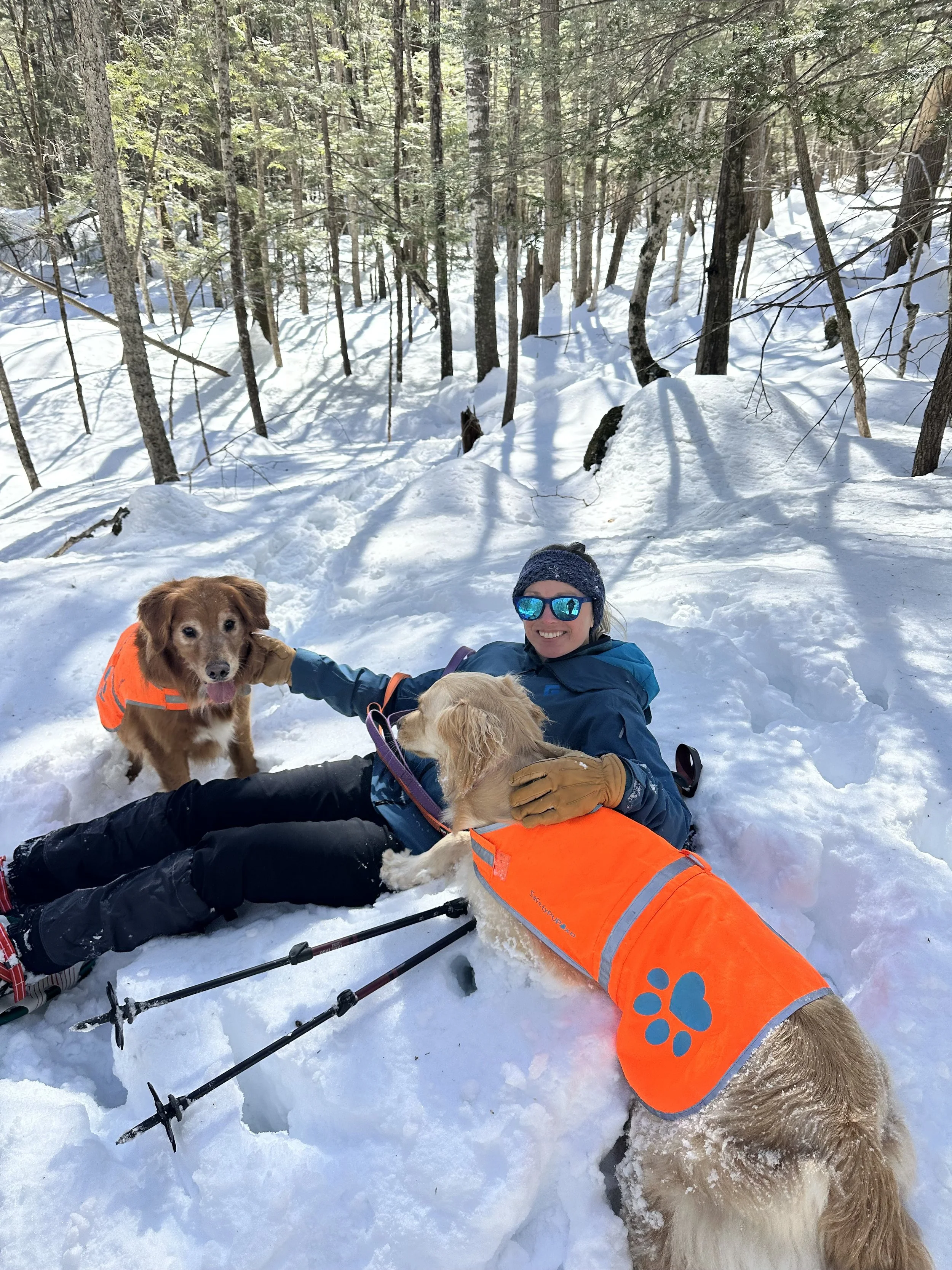 A woman with sunglasses sitting in the snow in a forest, smiling and holding two golden retrievers, while snow-covered trees surround them.