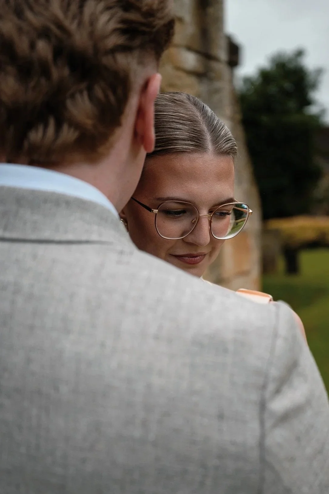 A close-up of a man and woman showing an intimate moment, with the man facing away and the woman with glasses looking down, outdoors near a stone wall and greenery.