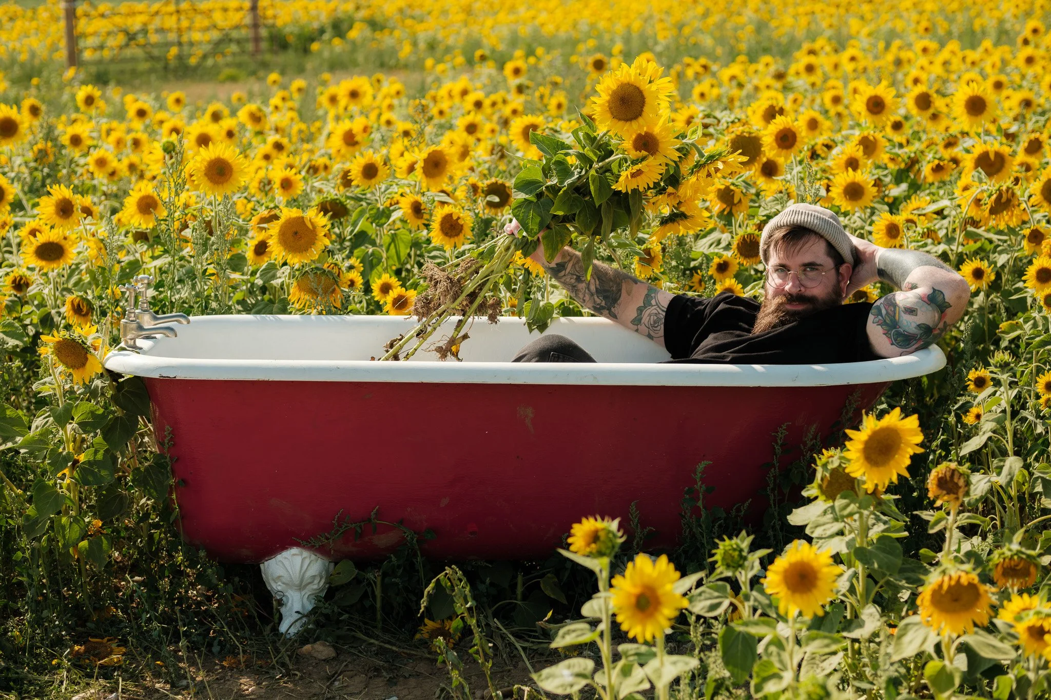 A man with tattoos lying in a vintage bathtub, holding a bouquet of sunflowers in a field of sunflowers.