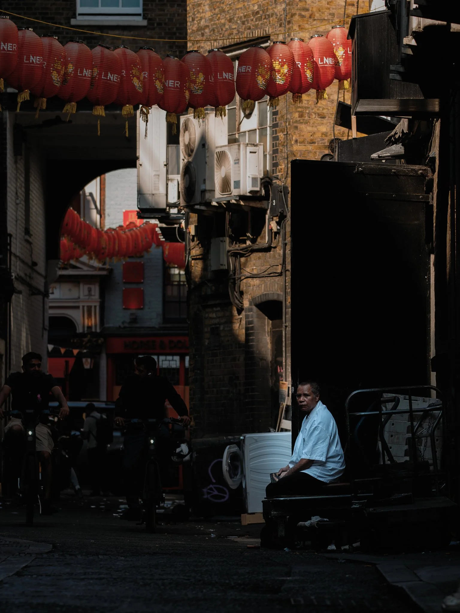 A street scene with red Chinese lanterns hanging across the street, brick buildings, and air conditioning units. Several people are on motorcycles, and a man with a white shirt is sitting on a cart or bench in the foreground. The scene is dimly lit w