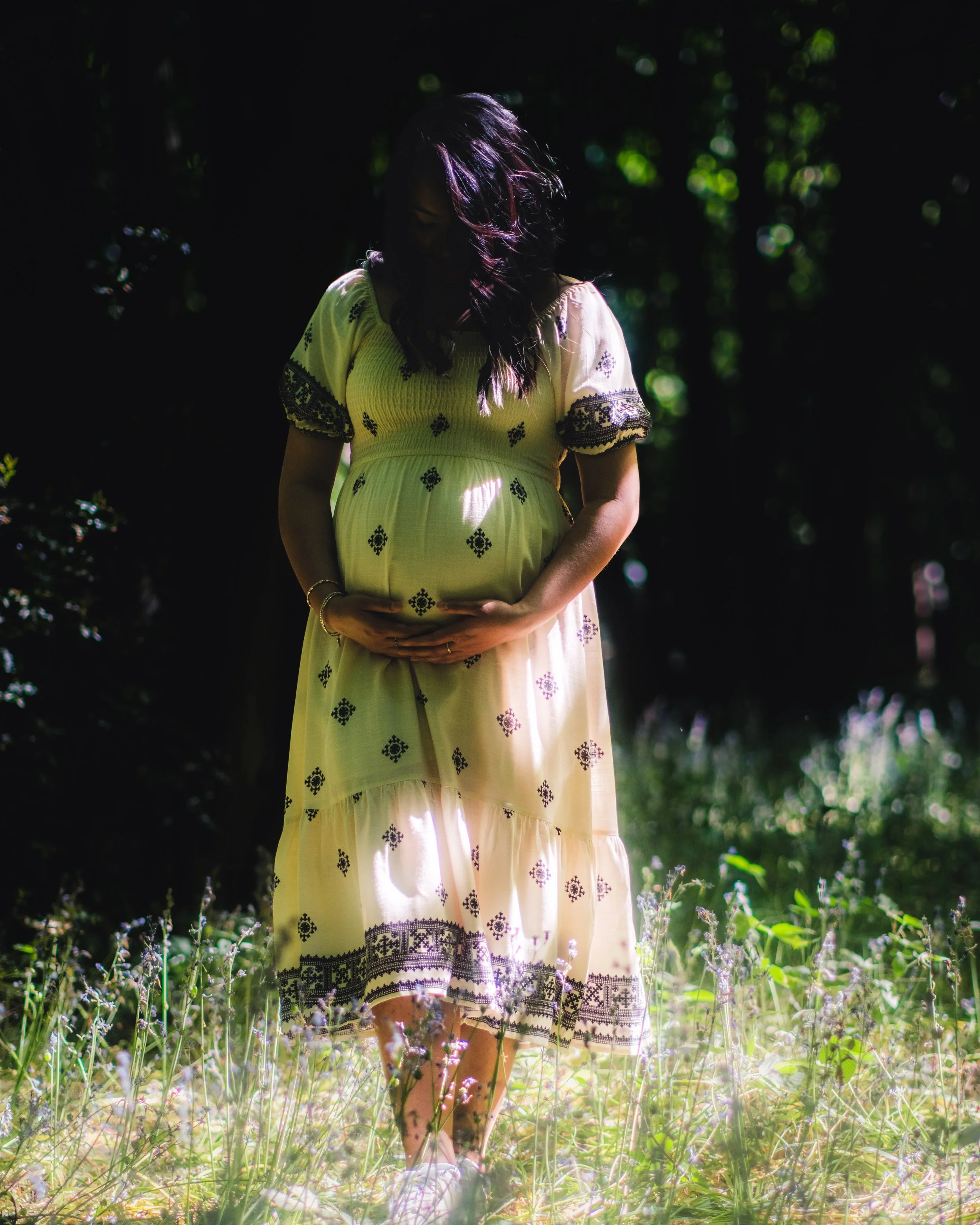 A woman in a yellow dress with black embroidery is standing in a garden, holding her pregnant belly, with sunlight and dark trees in the background.