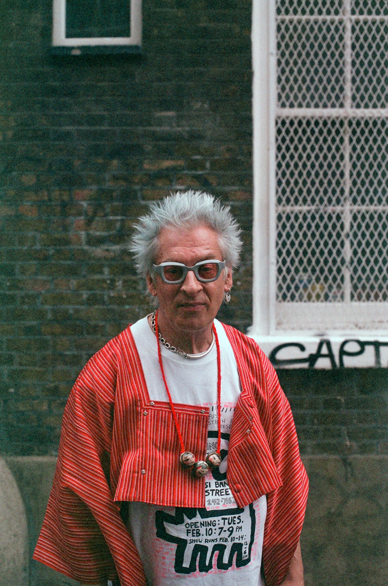 An older person with white spiky hair and sunglasses, wearing a red striped jacket over a graphic t-shirt, standing outdoors in front of a dark brick wall and a white metal gate.