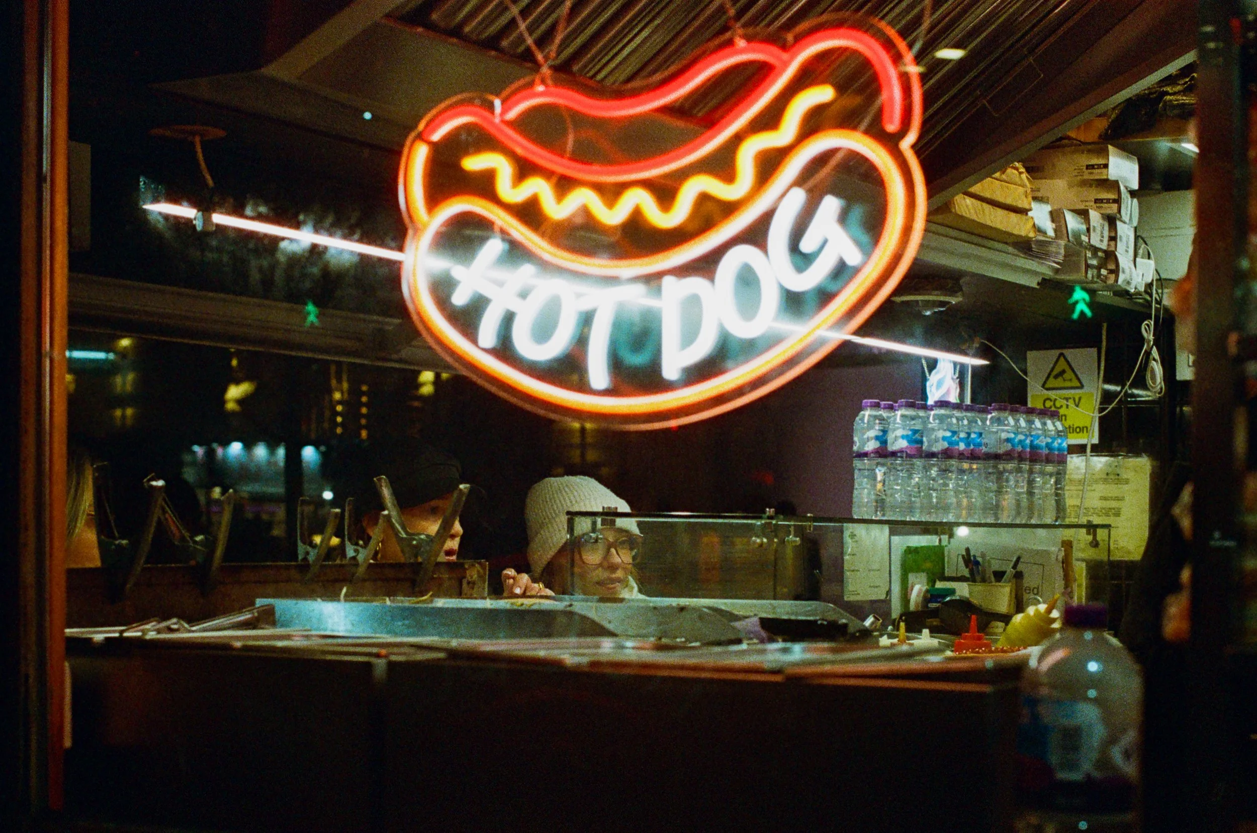 Neon sign with a hot dog illustration and the words "Hot Dog" inside a heart shape, viewed through a window at night.