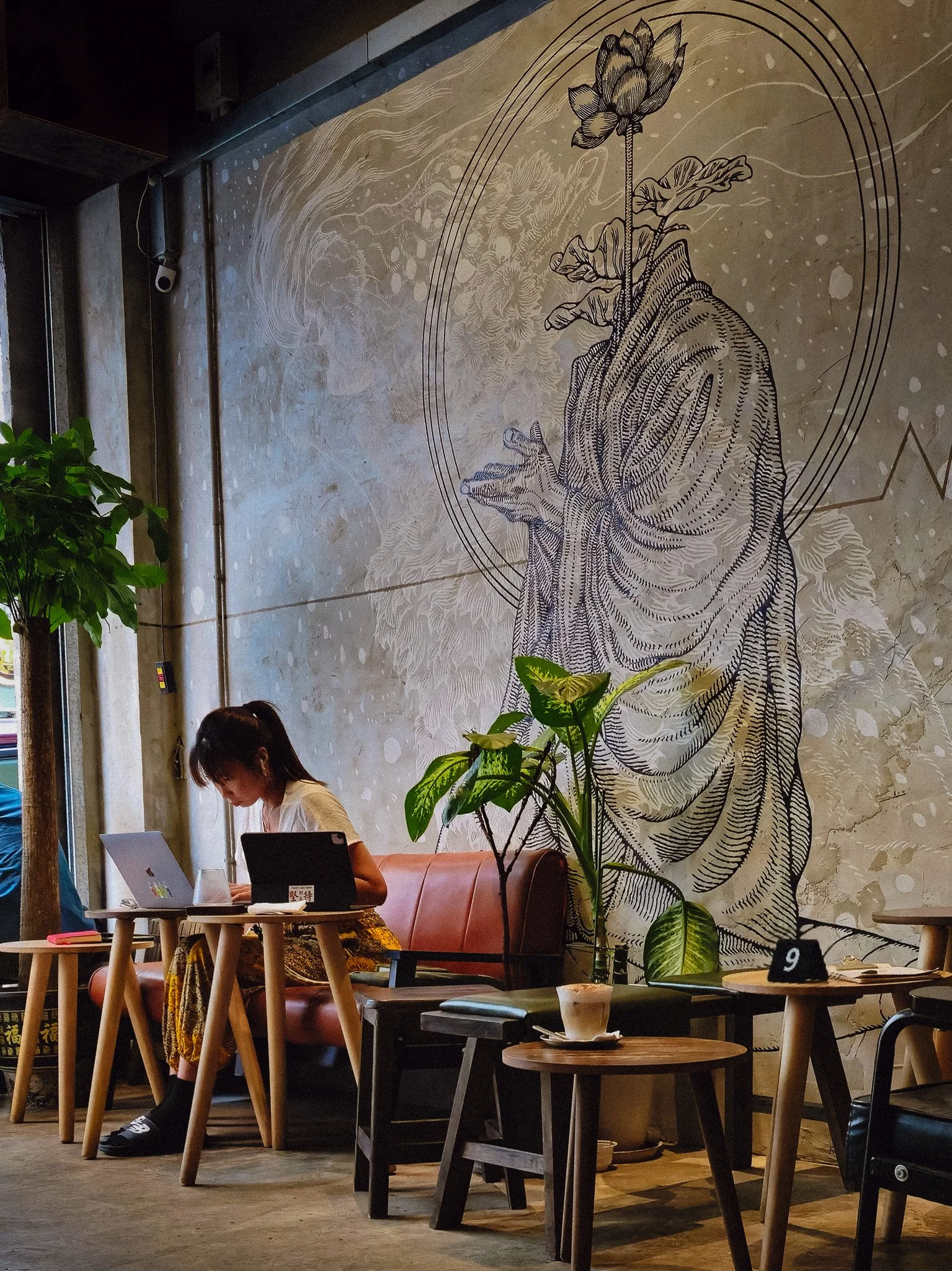 Interior of a cafe with a woman working on a laptop, decorated with a large wall art of a hand holding a flower, green plants, wooden tables, and chairs.