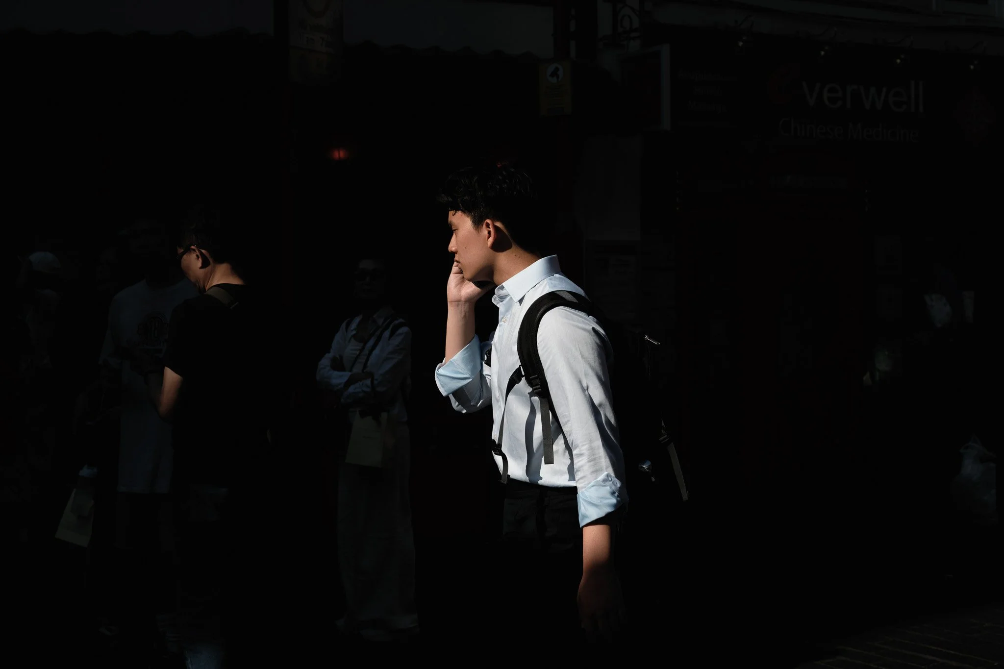 Young man with black hair, wearing a white shirt and carrying a backpack, talking on his cellphone in a busy area with other people in the background.