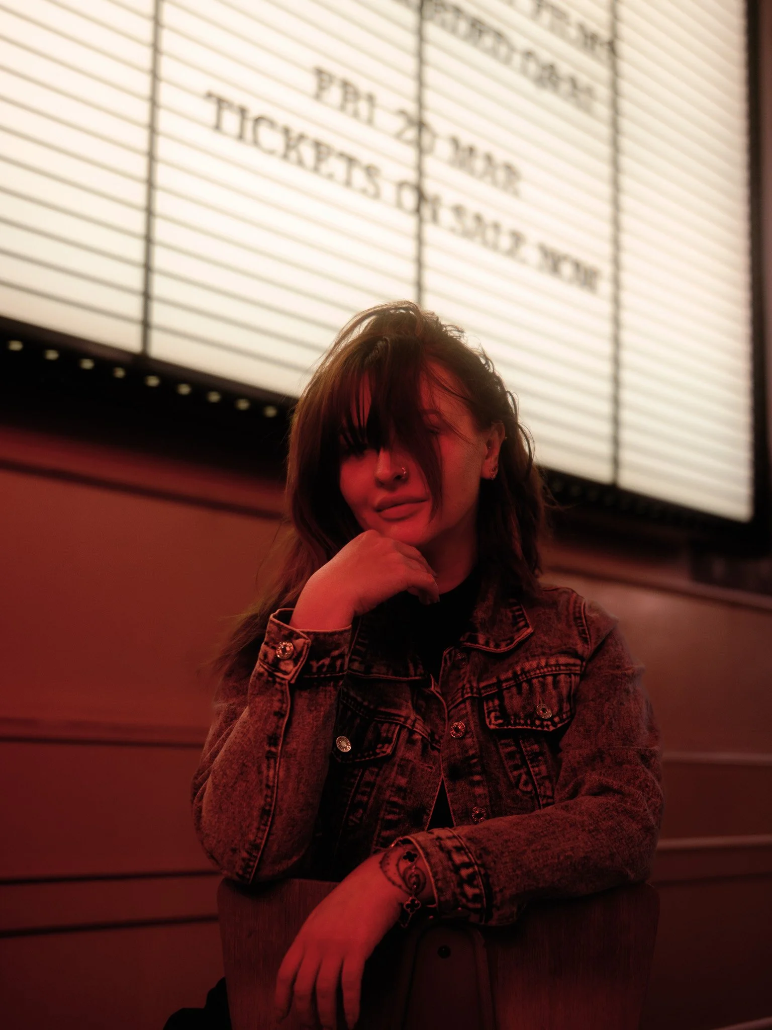 A woman with brown hair and a nose ring, wearing a denim jacket and black shirt, sitting at a table with her chin resting on her hand, in front of a lit-up movie theater marquee sign.