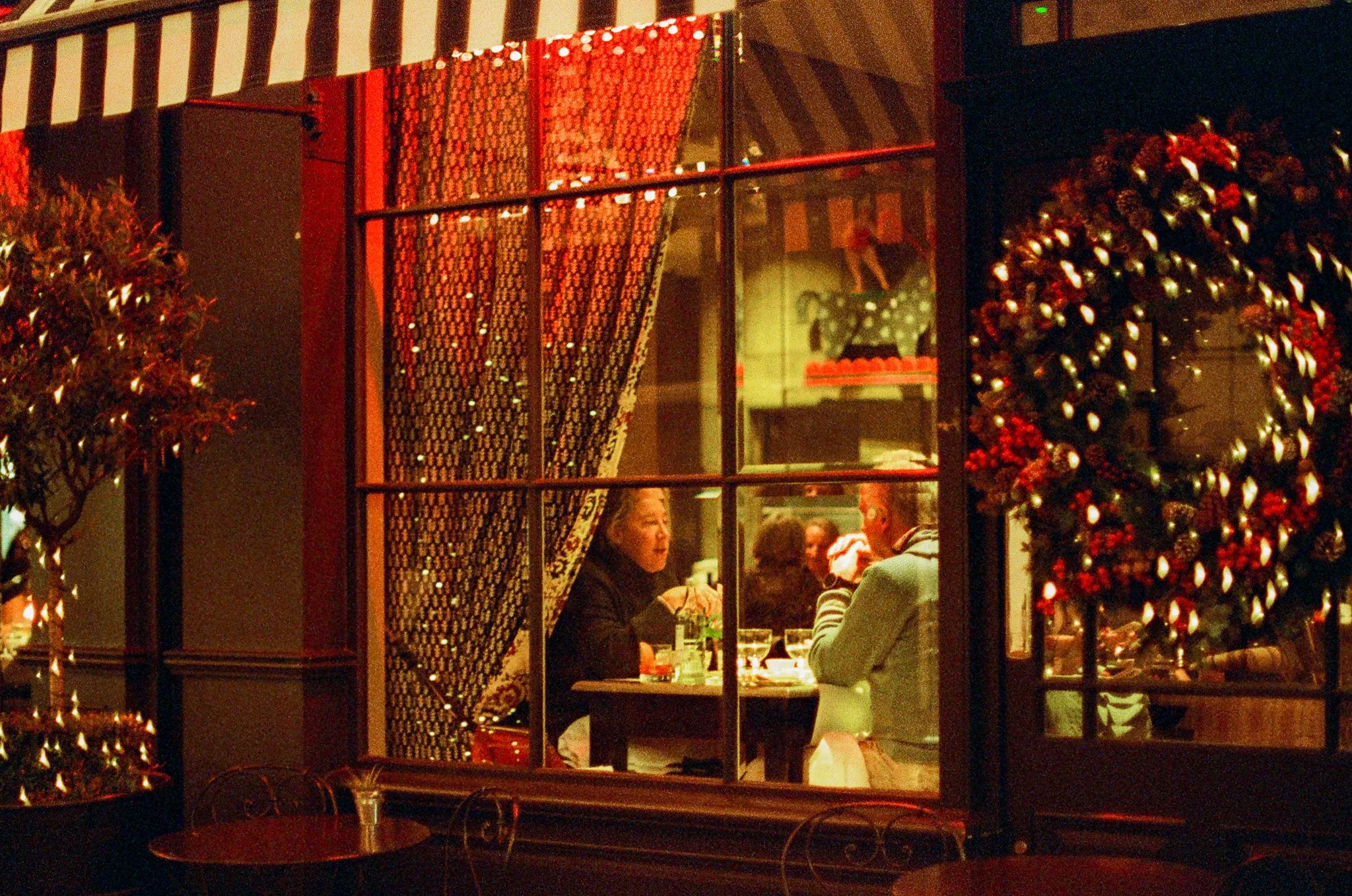 A scene viewed through a large window at night, showing people sitting at a table inside a restaurant decorated with Christmas ornaments and warm lighting. Outside the window, a Christmas tree with red and gold ornaments is visible. The reflection in