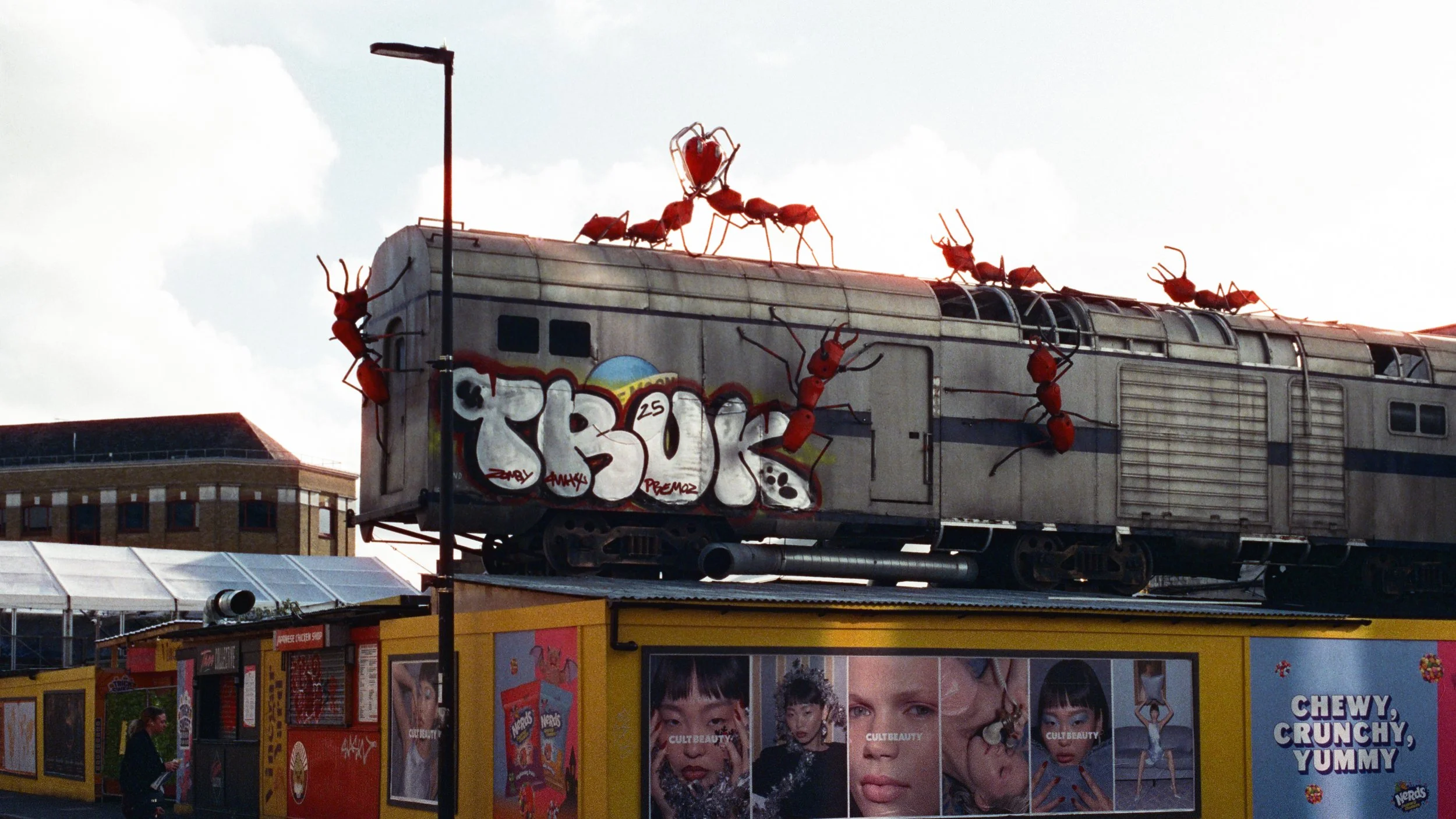 A train car with large red ant sculptures on top, some carrying hearts, and graffiti spelling 'TRUTH' on its side, above a yellow building with advertisements and posters.