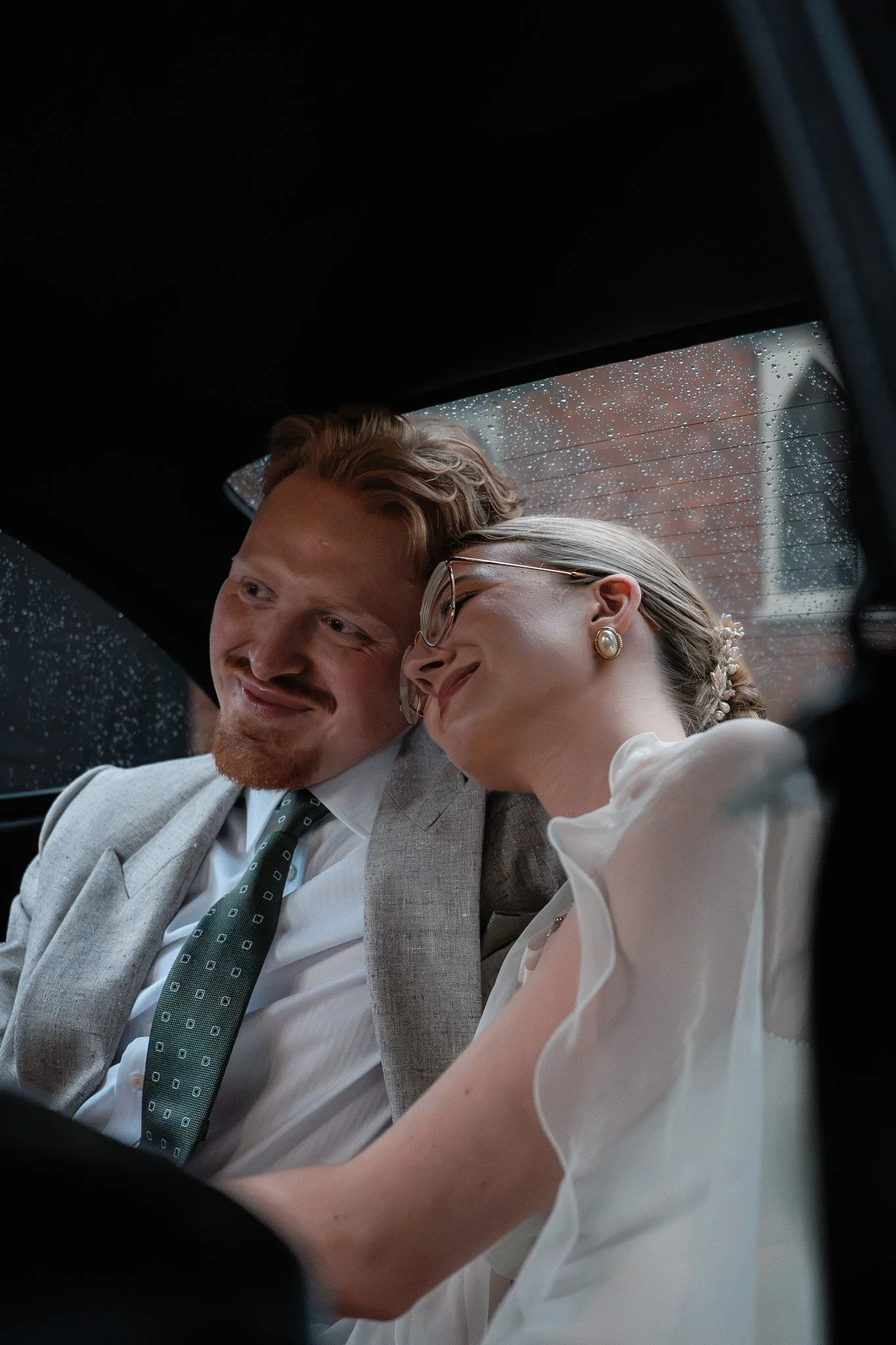Close-up of a newlywed couple sitting inside a car during a rainy day, smiling and leaning on each other.