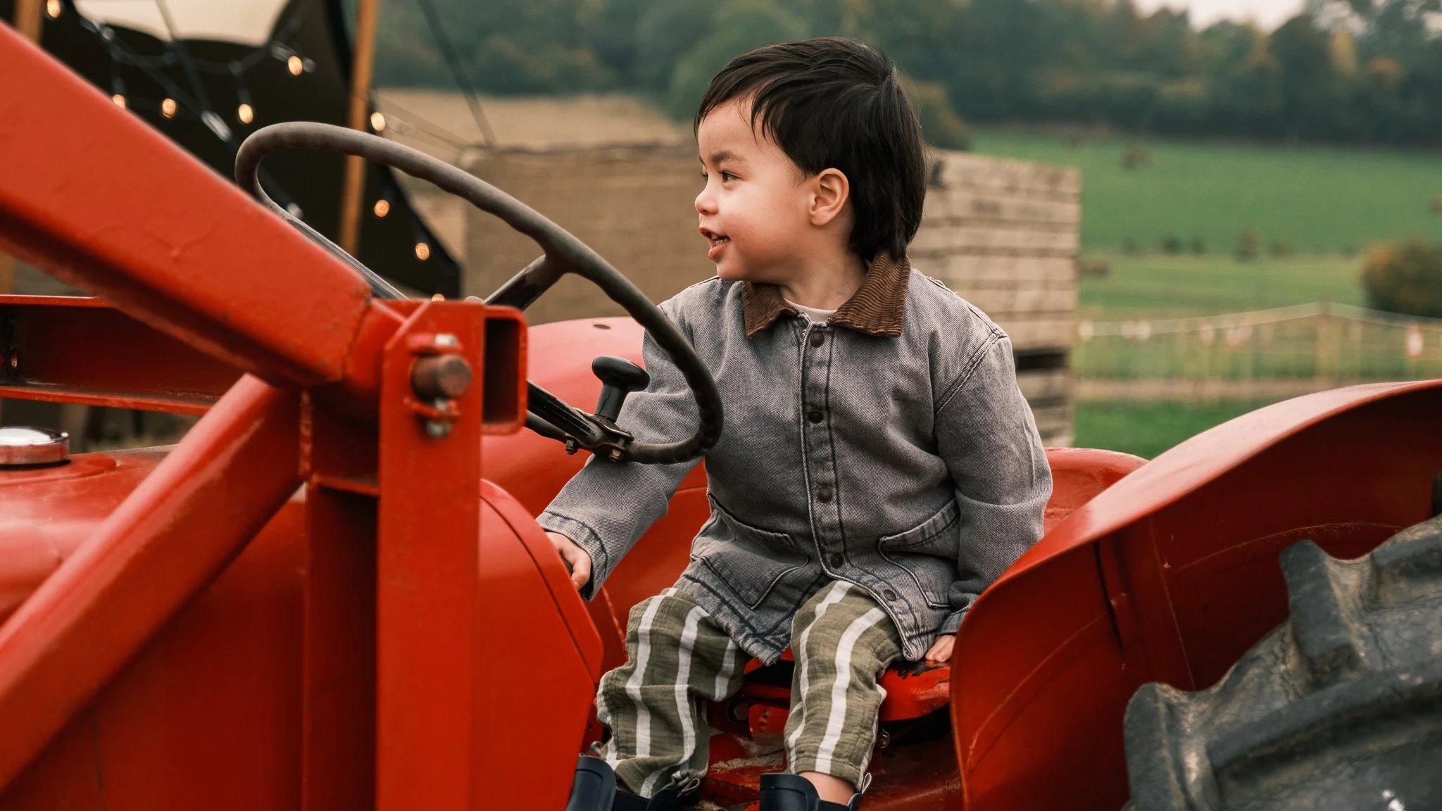 A young boy sitting on a red tractor outdoors with a field and trees in the background.