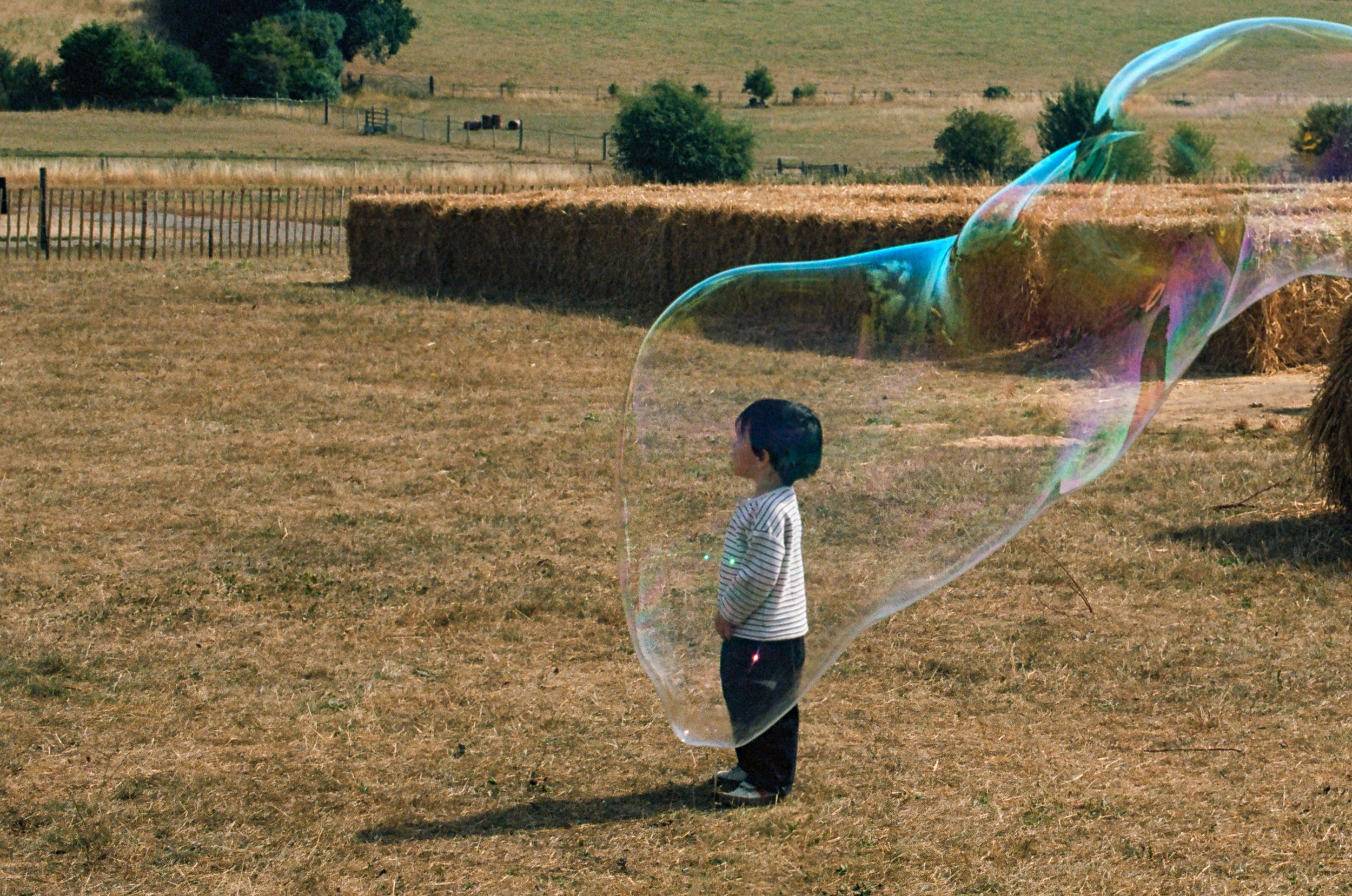 A young boy standing in a field with dry grass, playing with large soap bubbles during daytime. In the background, there are hay bales, trees, and a wooden fence.
