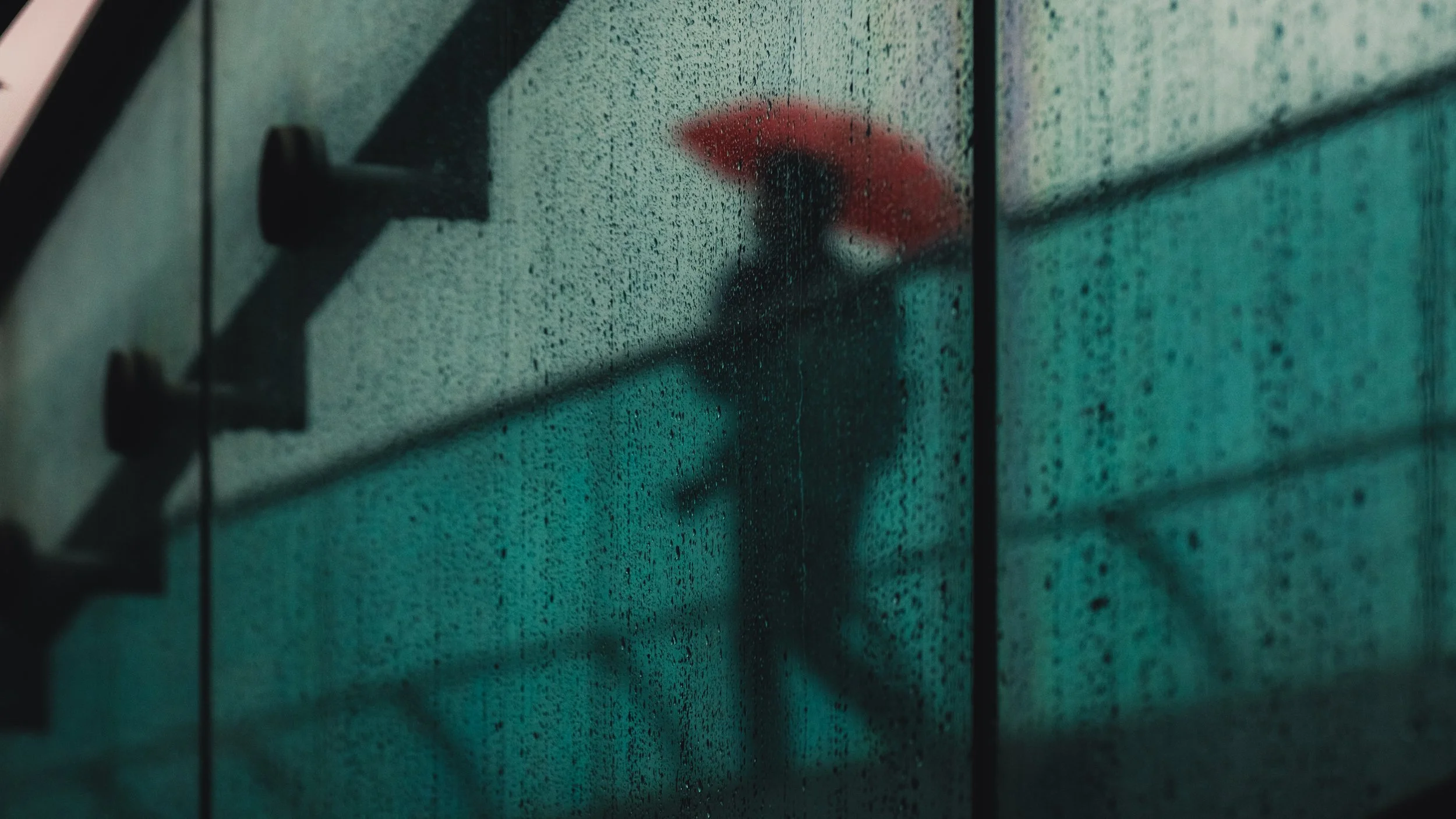 A blurry silhouette of a person holding an umbrella, reflected on a rain-covered window.