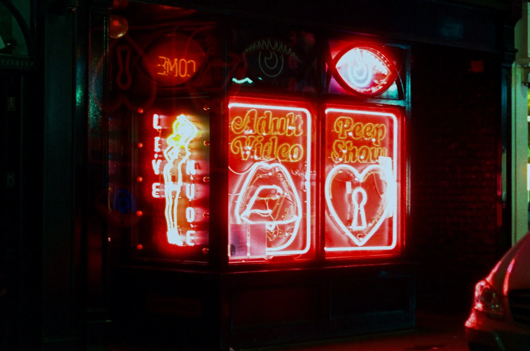 A cluster of red neon signs displays advertisements for a coffee shop, including words like 'Love,' 'Café,' 'Coffee,' 'Admit Video,' and 'Peep Show.' The signs include images of lips and a heart, creating a vibrant, colorful glow in a dark setting.