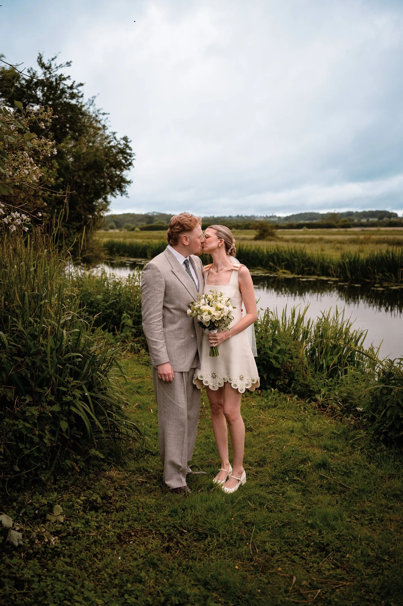 A couple is kissing outdoors by a river with grassy fields and trees in the background, during overcast weather, with the woman holding a bouquet of flowers.