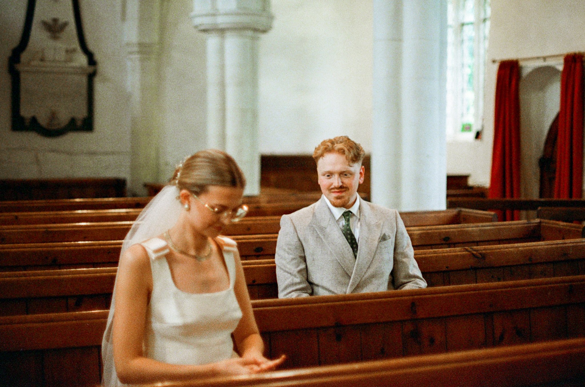 A man in a gray suit and a woman in a white dress are sitting on wooden pews inside a church, with red curtains and stained glass windows in the background.