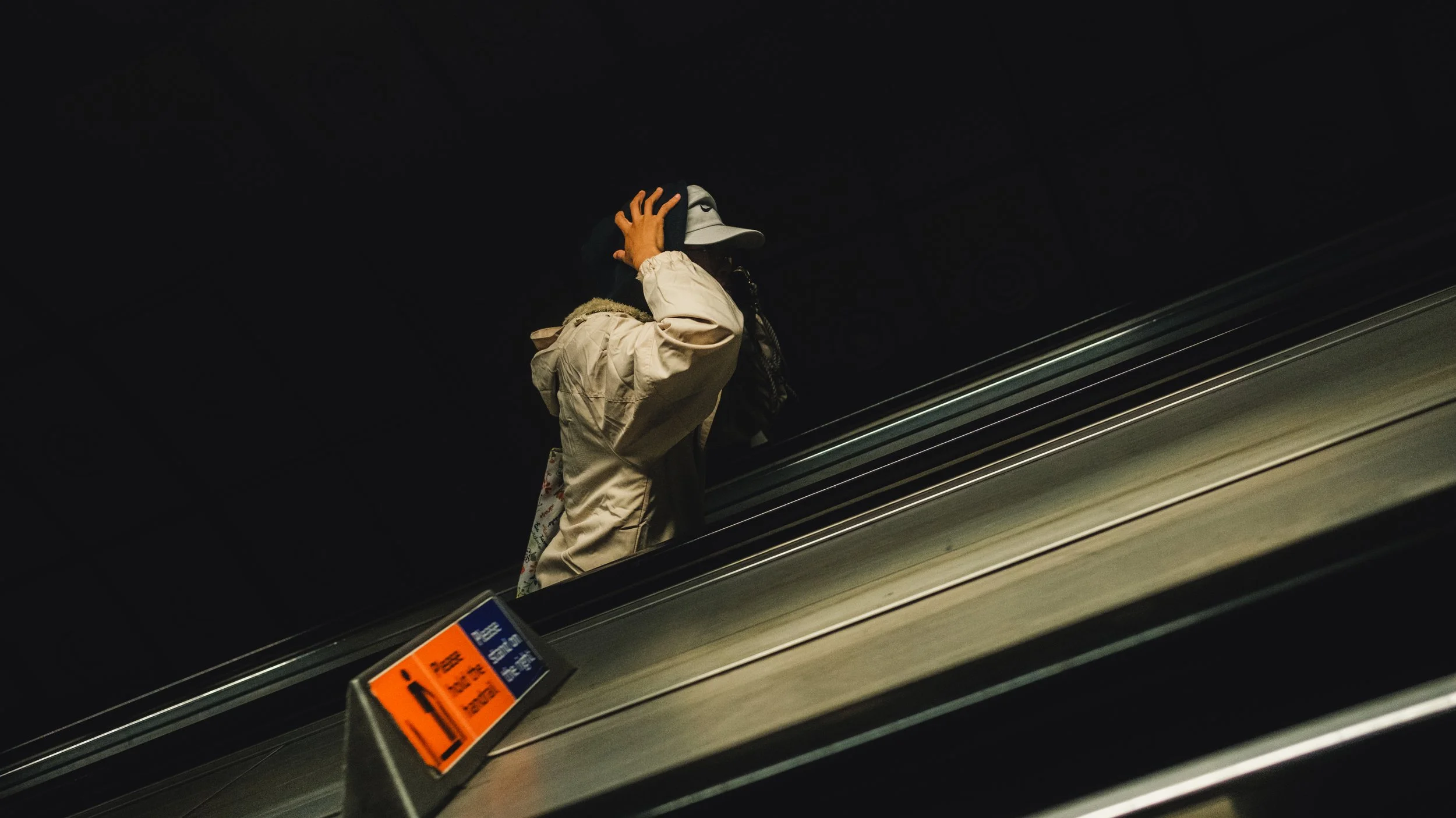 Person wearing a beige jacket and a face mask, holding a hat on an escalator in a dark environment.