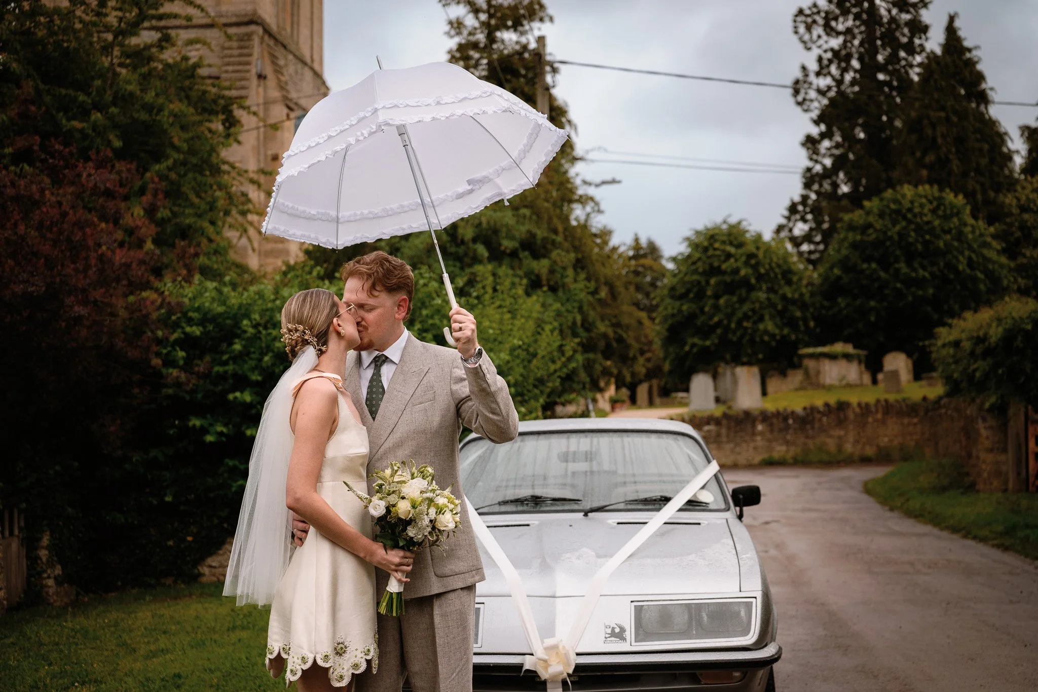 A bride and groom sharing a kiss under a white umbrella outdoors on a cloudy day, with a white car decorated with ribbon in the background, and trees and buildings in the distance.