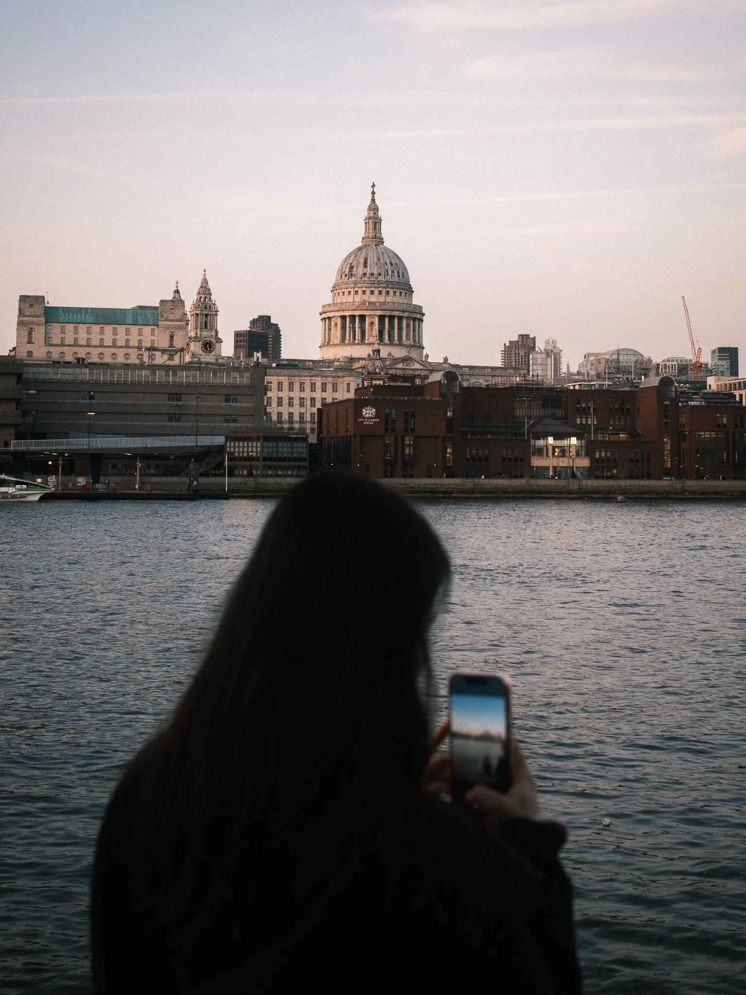 View of St. Paul's Cathedral in London seen from across the river Thames during sunset, with a person's hand in the foreground holding a phone and a finger pointing upwards.