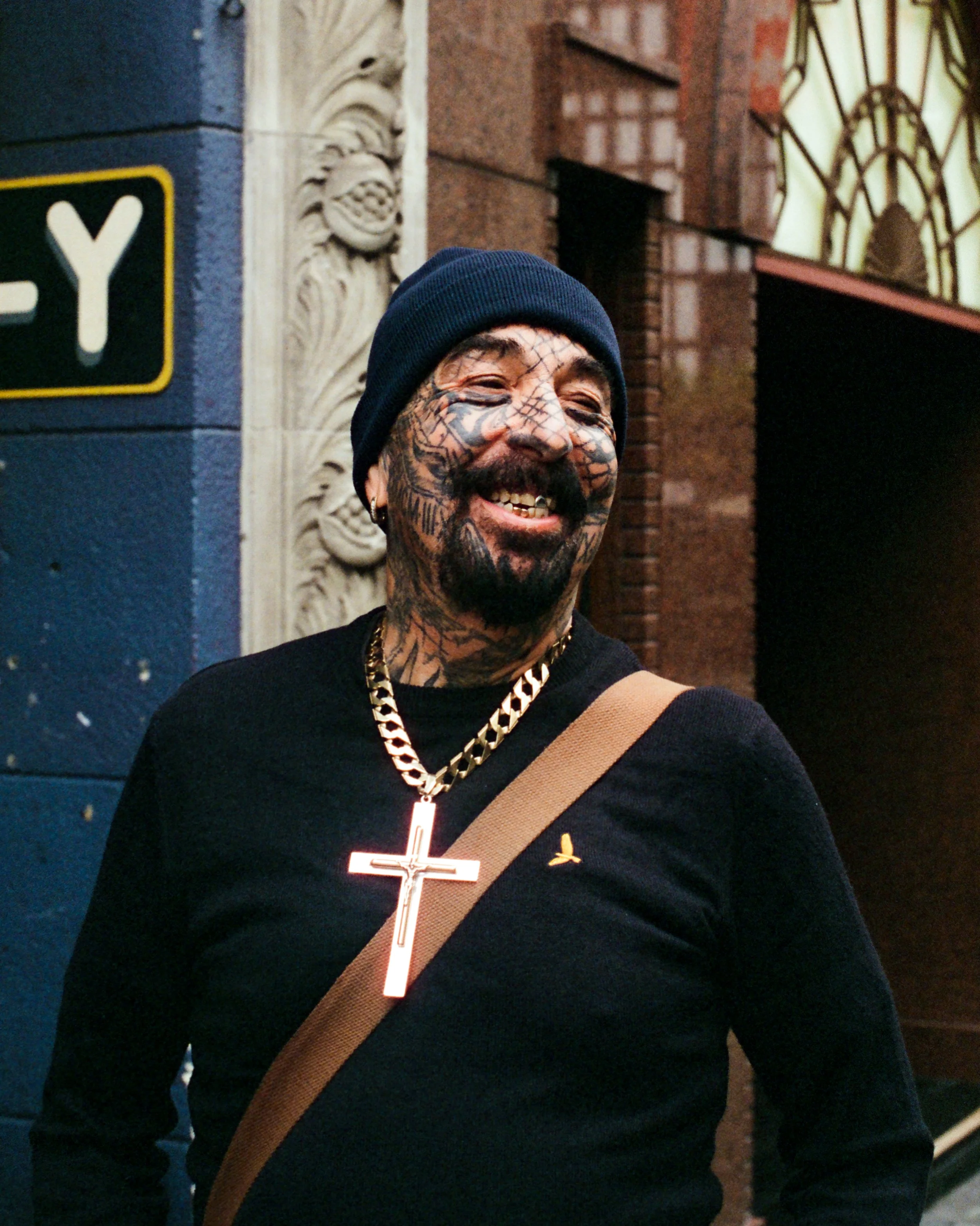A smiling man with a mustache and tattoos on his face, wearing a black beanie, a gold chain, and a black shirt with a gold ribbon emblem. He has a cross necklace and a tan crossbody bag, standing against a brick and decorative stone wall with a stree