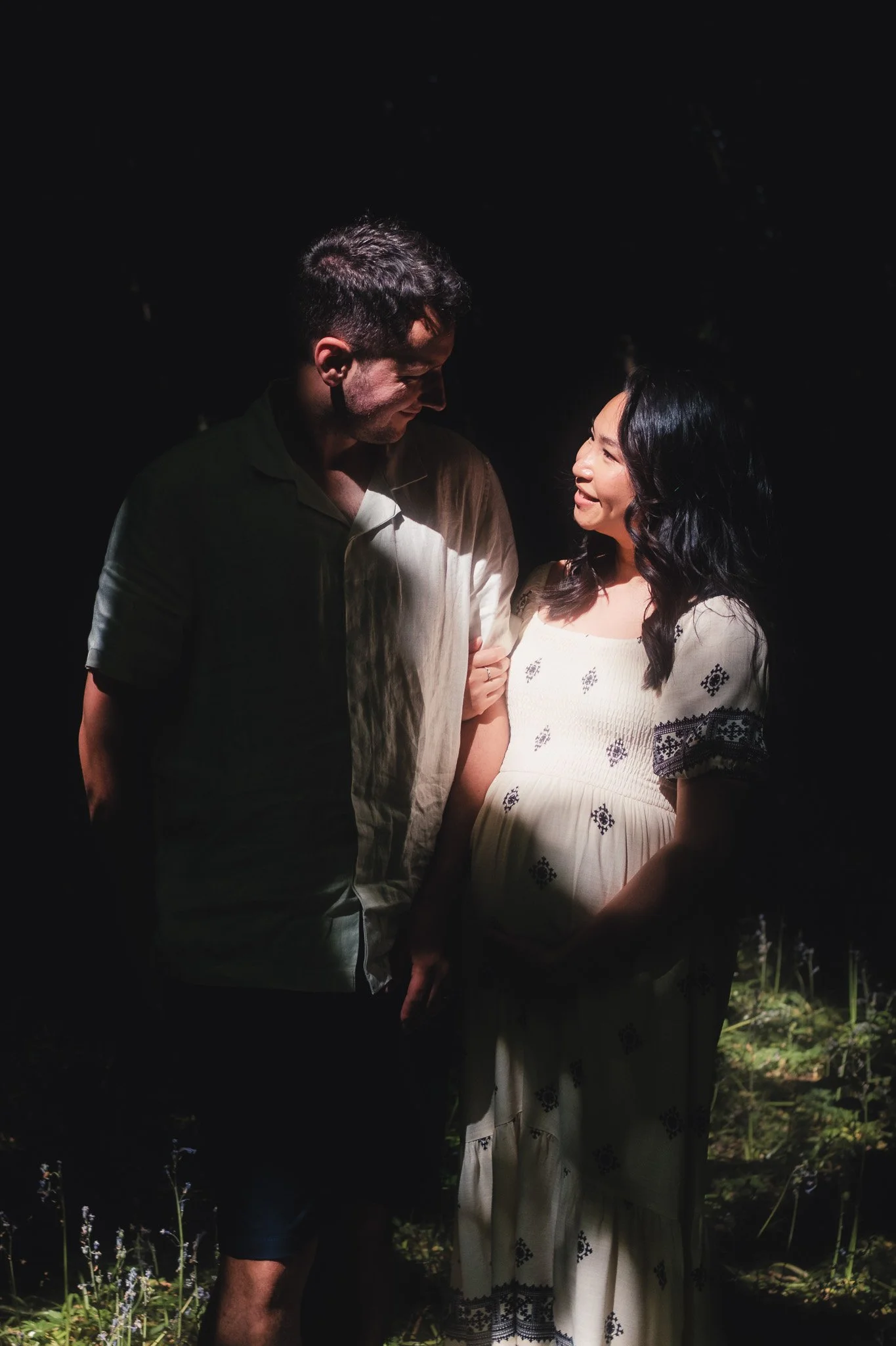 A man and a woman are sharing a moment of happiness outdoors, with the woman being pregnant. The woman is smiling and looking at the man, who is looking down at her, both illuminated by soft light, against a dark natural background.