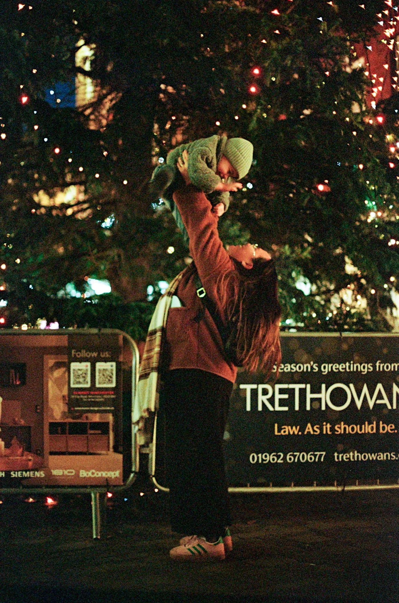 A person lifts a small child in front of a decorated Christmas tree with colorful lights, at nighttime. The person is wearing dark pants, a brown jacket, sneakers, and has long hair. The child is wearing a gray hoodie, cap, and jeans. There is a sign
