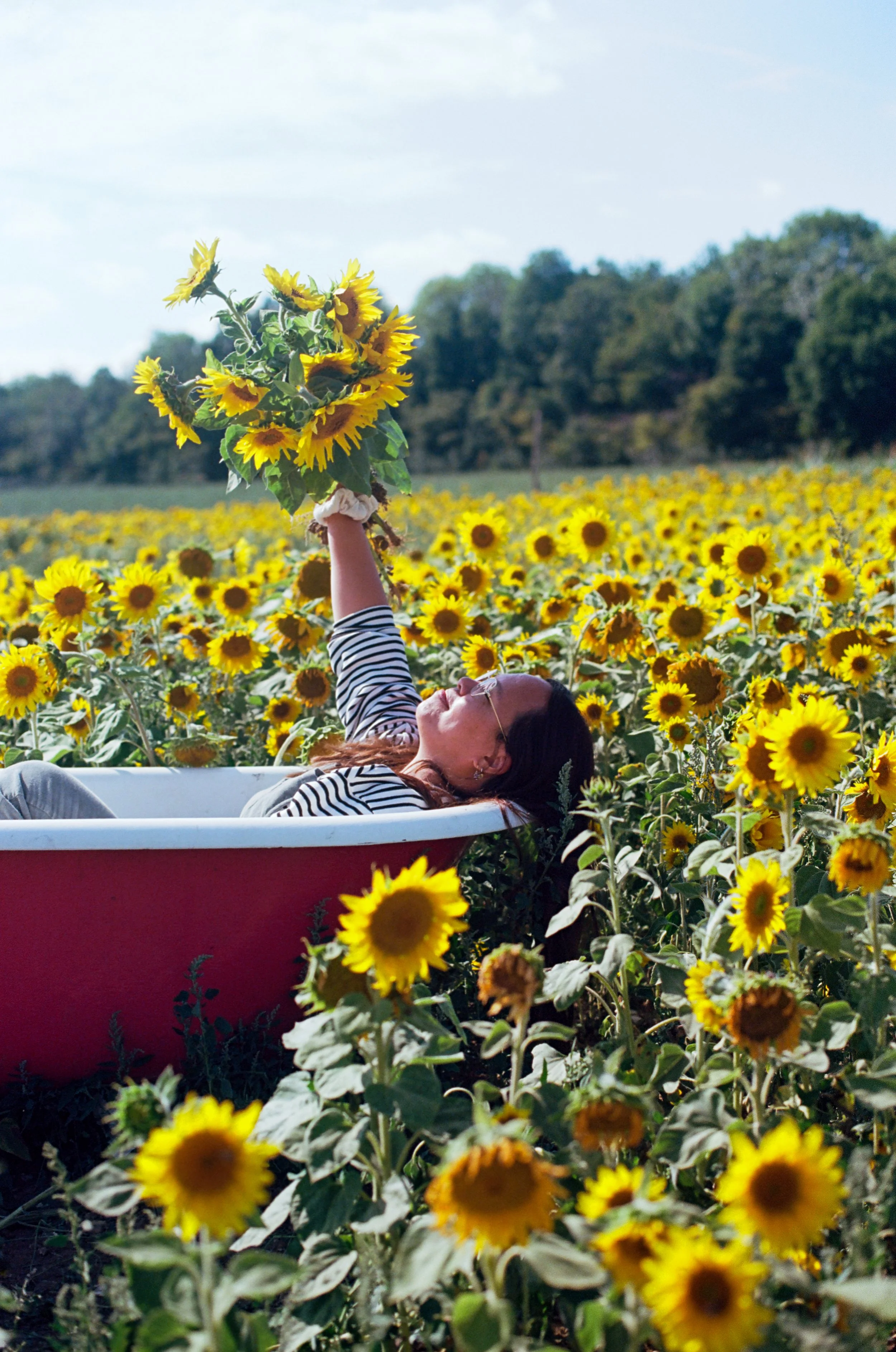 A woman lying in a red boat in a sunflower field, holding a bouquet of sunflowers, with a forested background and a blue sky.