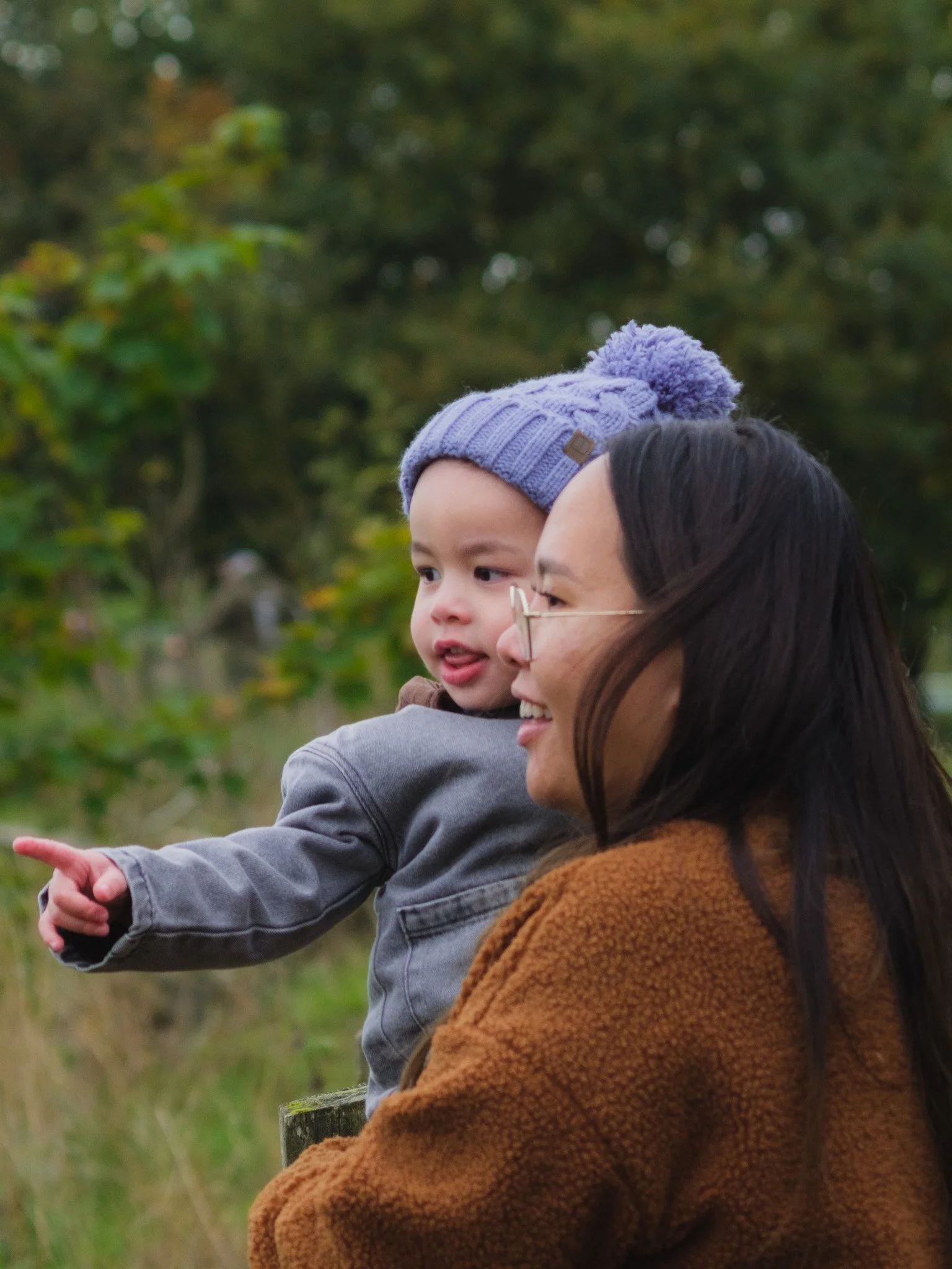A woman and a young child outdoors, with the child pointing at something in the distance. The woman has long dark hair and is wearing glasses and a brown coat. The child is wearing a purple knit hat with a pom-pom and a gray jacket. The background fe